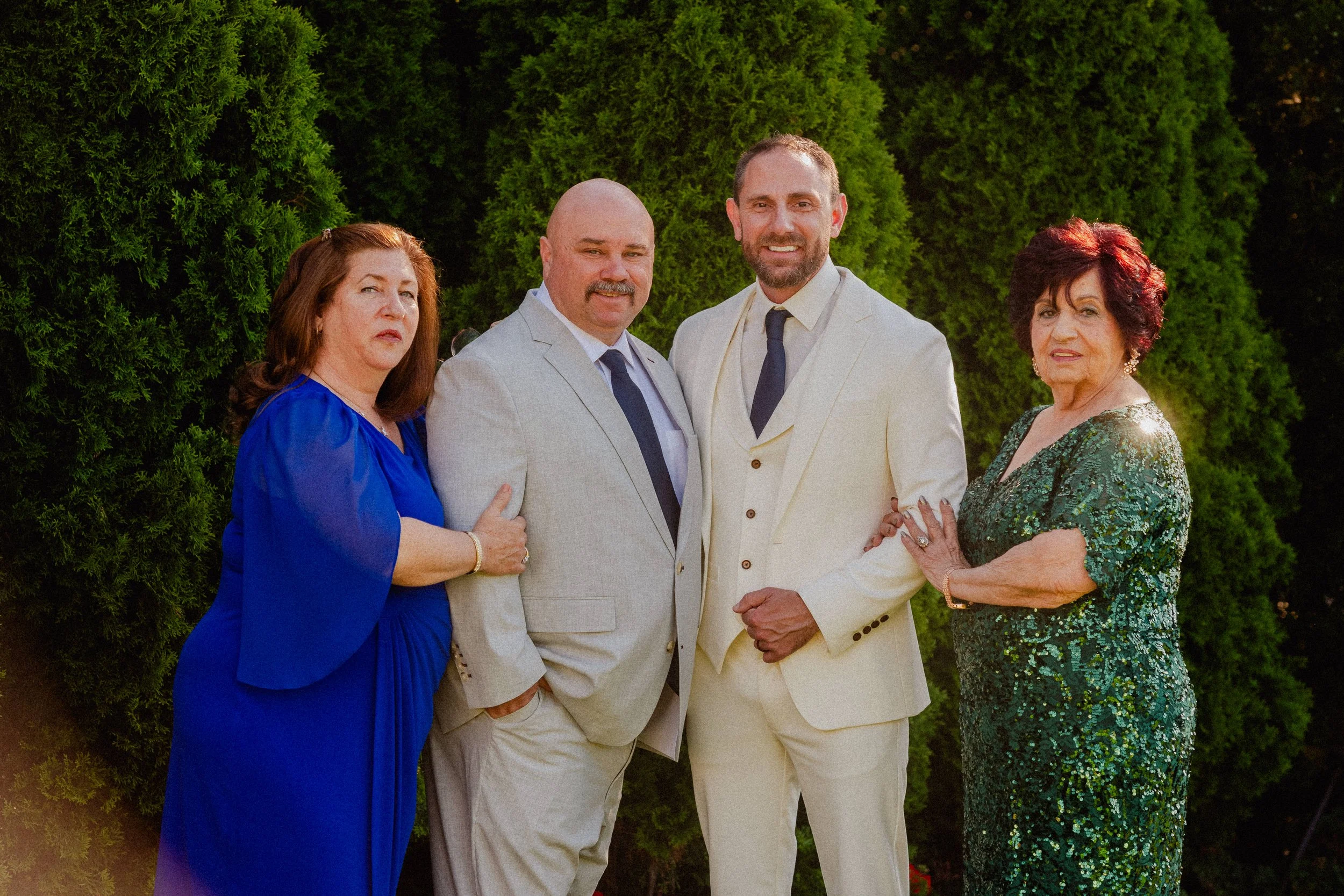 A group of four adults, two women and two men, standing outdoors in front of lush green bushes. The women are dressed in elegant gowns, one in blue and the other in green, while the men are wearing suits, one in grey and the other in cream. They are 
