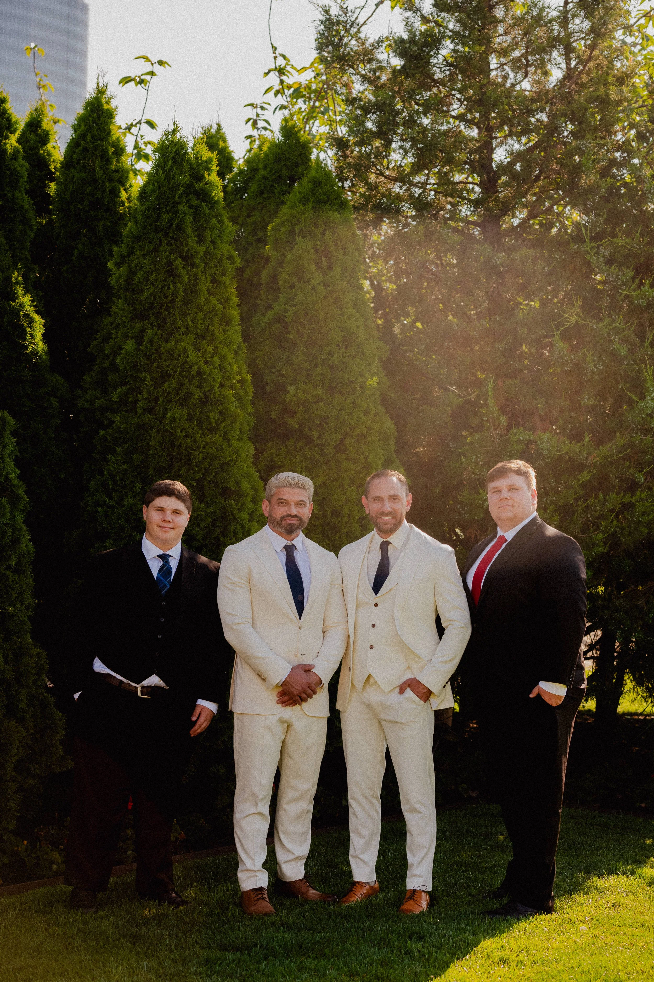 Four men in suits standing outdoors on grass, with tall greenery and trees behind them, during daylight.