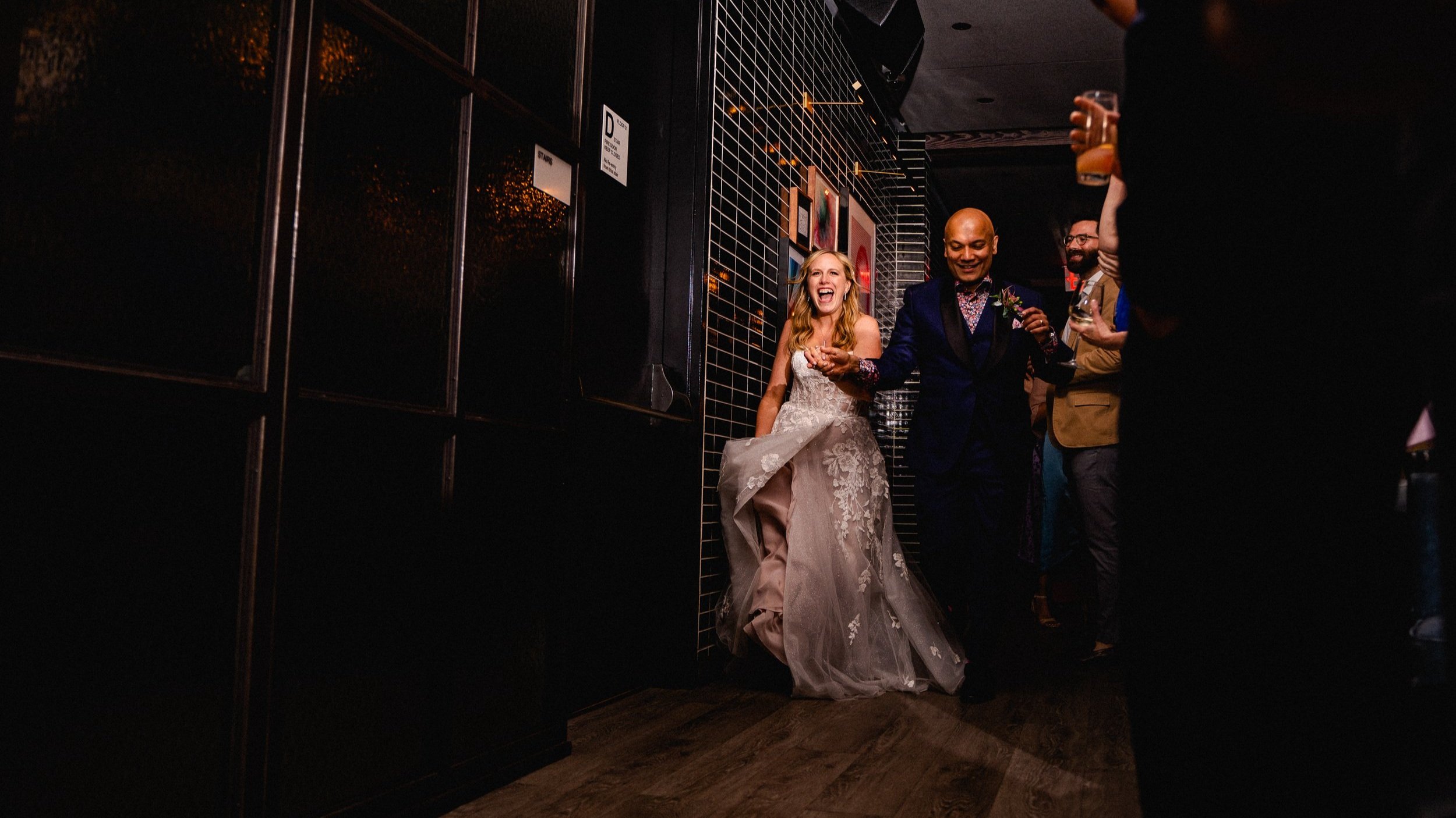 Bride and groom walking in a dimly lit venue, smiling and holding hands, with friends enjoying the celebration around them.