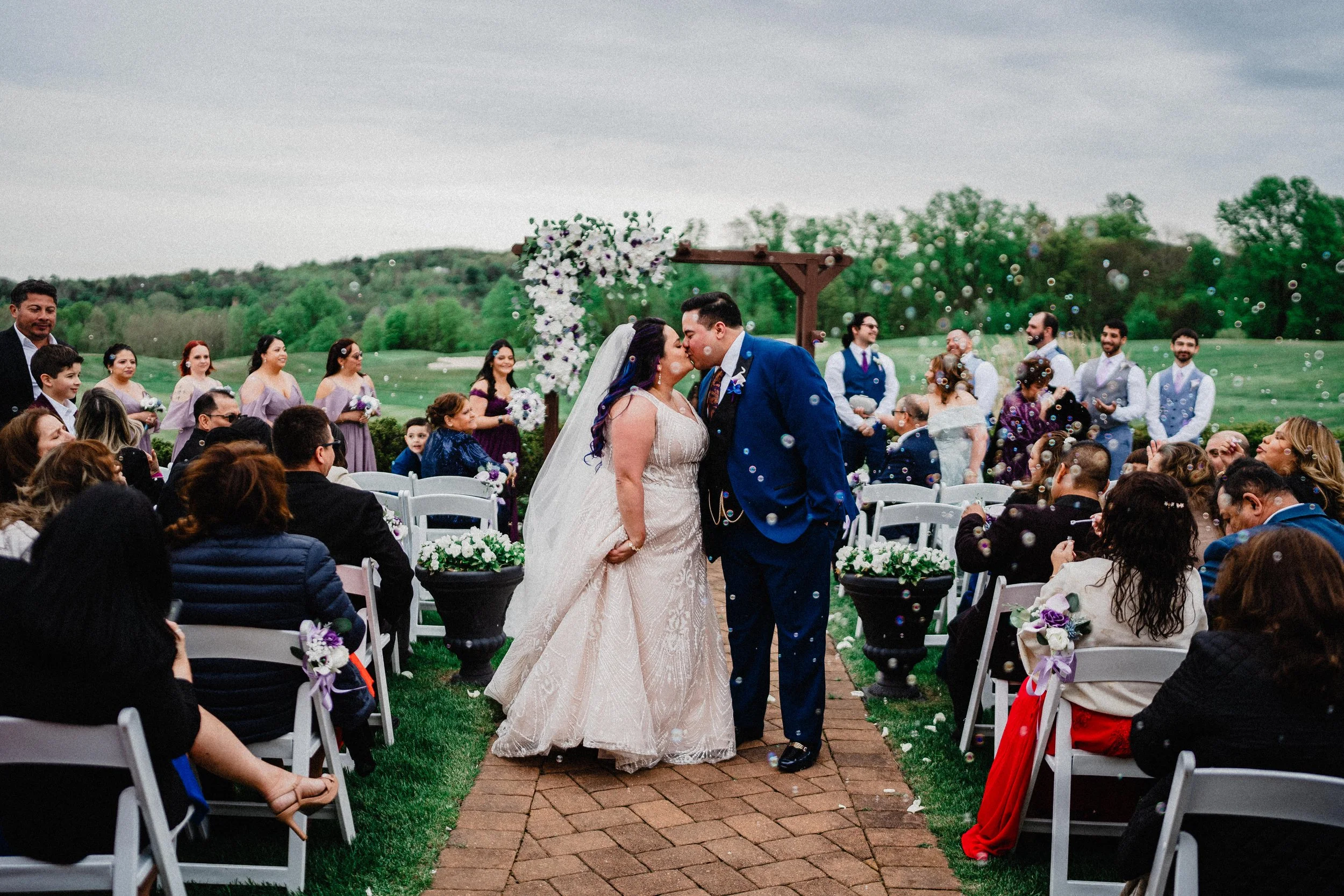 A bride and groom kiss during their outdoor wedding ceremony, surrounded by friends and family on a green lawn with a cloudy sky in the background.