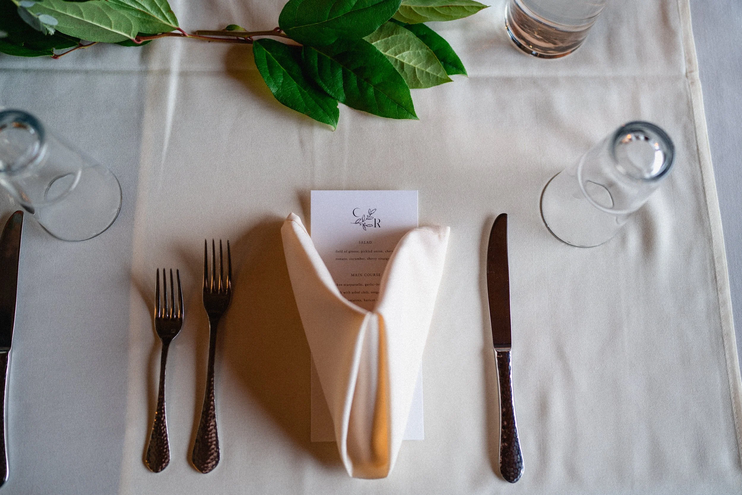 Elegant dining table setting with two forks, a knife, a napkin, a menu card, two empty glasses, and a green leafy branch on a white tablecloth.