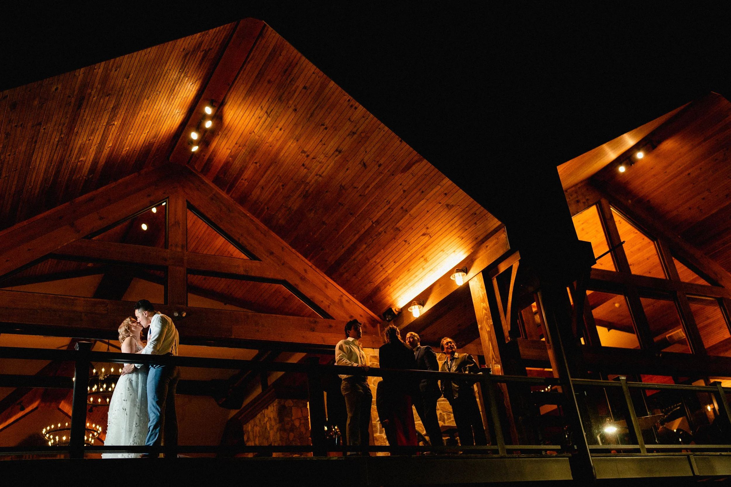 A couple dancing closely at a wedding reception during nighttime in a wood-paneled venue with warm lighting, while a group of people stands nearby on a balcony.