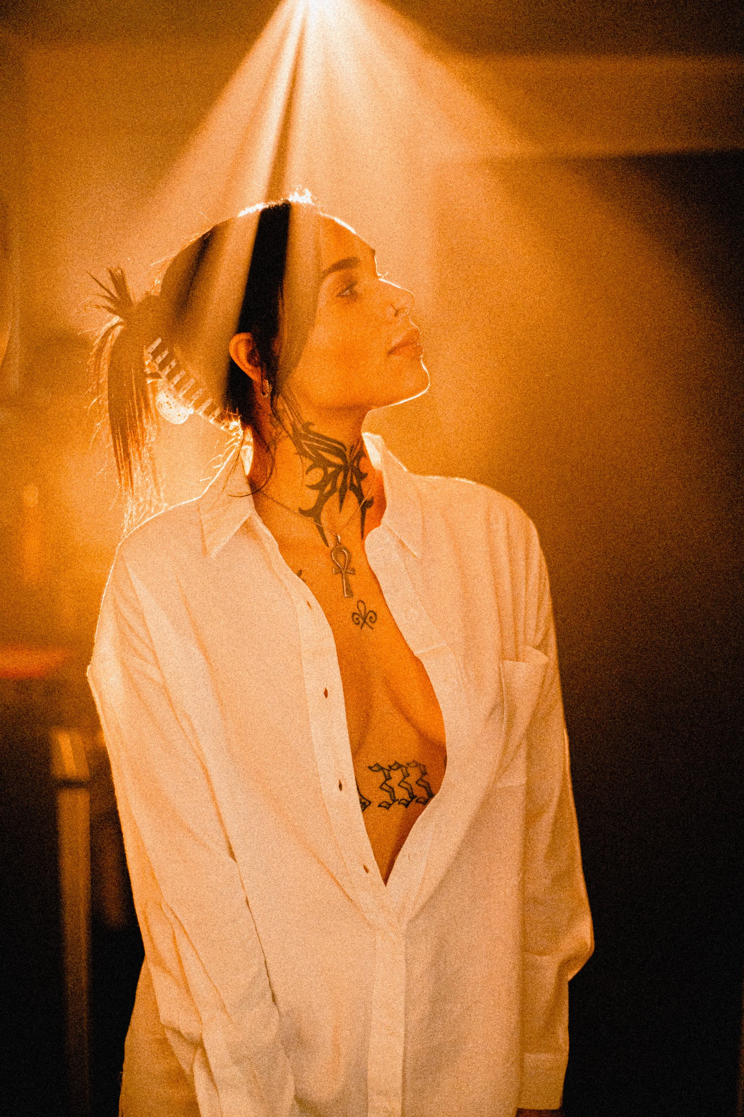 Commercial Photography- A woman with tattoos and a ponytail, wearing an open white shirt, stands in warm, dramatic lighting with her eyes closed, illuminated by a spotlight.