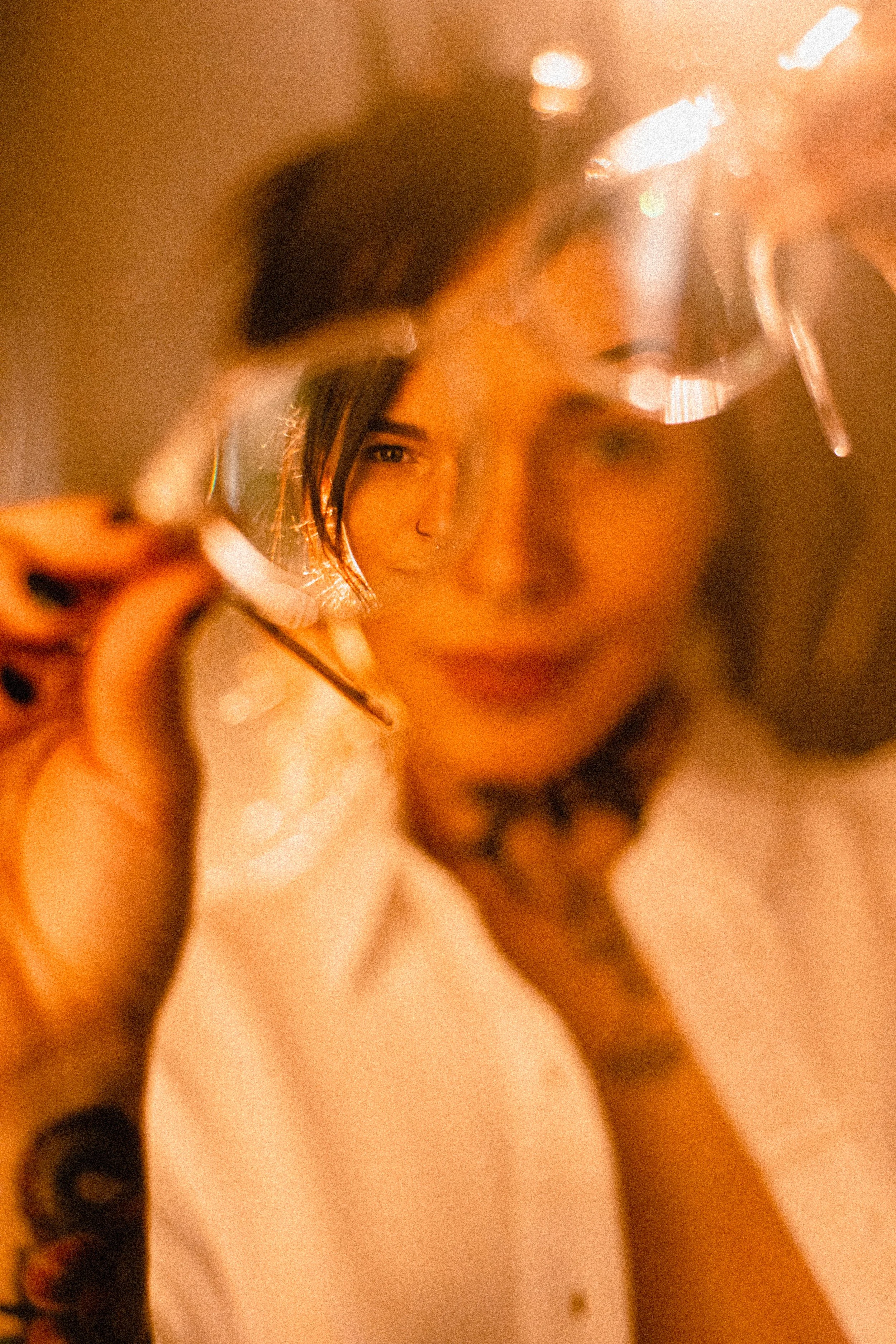 Commercial Photography- A woman in a white shirt is holding a glass with a drink, seen through the reflection of the glass she is holding, with a warm, orange lighting.