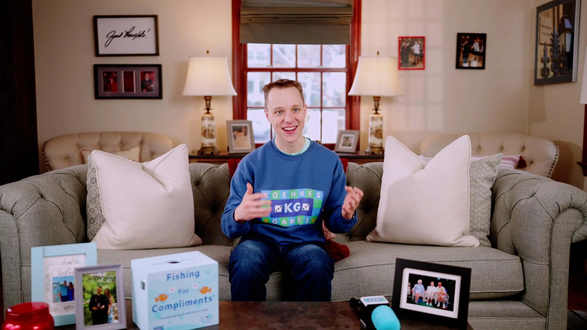 Video Production - A smiling man sitting on a beige couch in a cozy living room, surrounded by family photos, lamps, and decorative pillows.