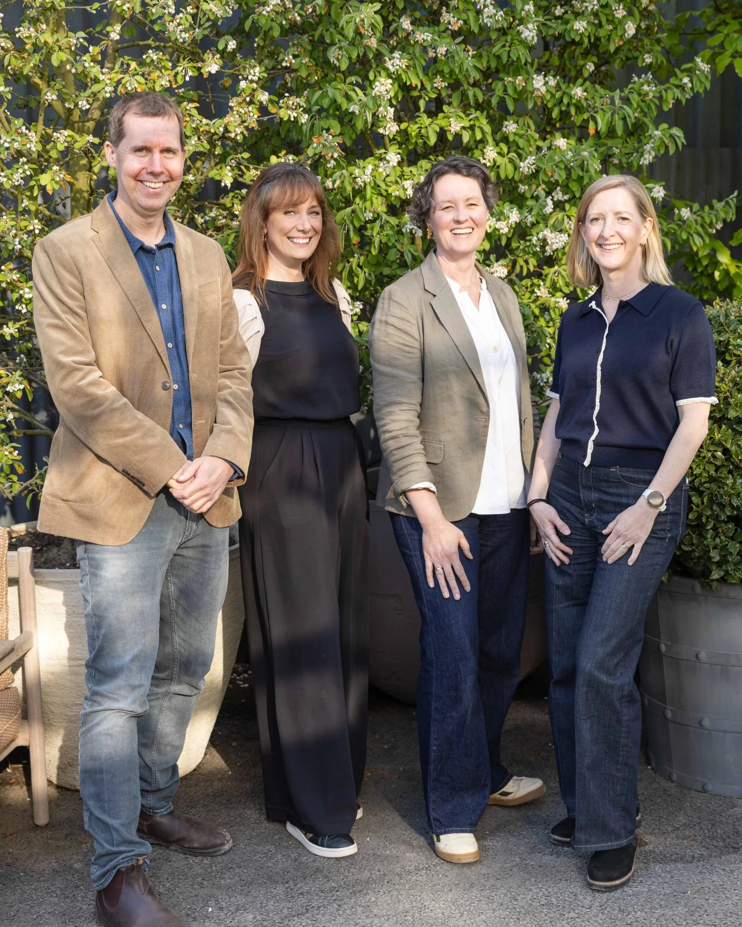 Group of four people standing outdoors in front of a green leafy shrub with white flowers, smiling and posing for the photo.