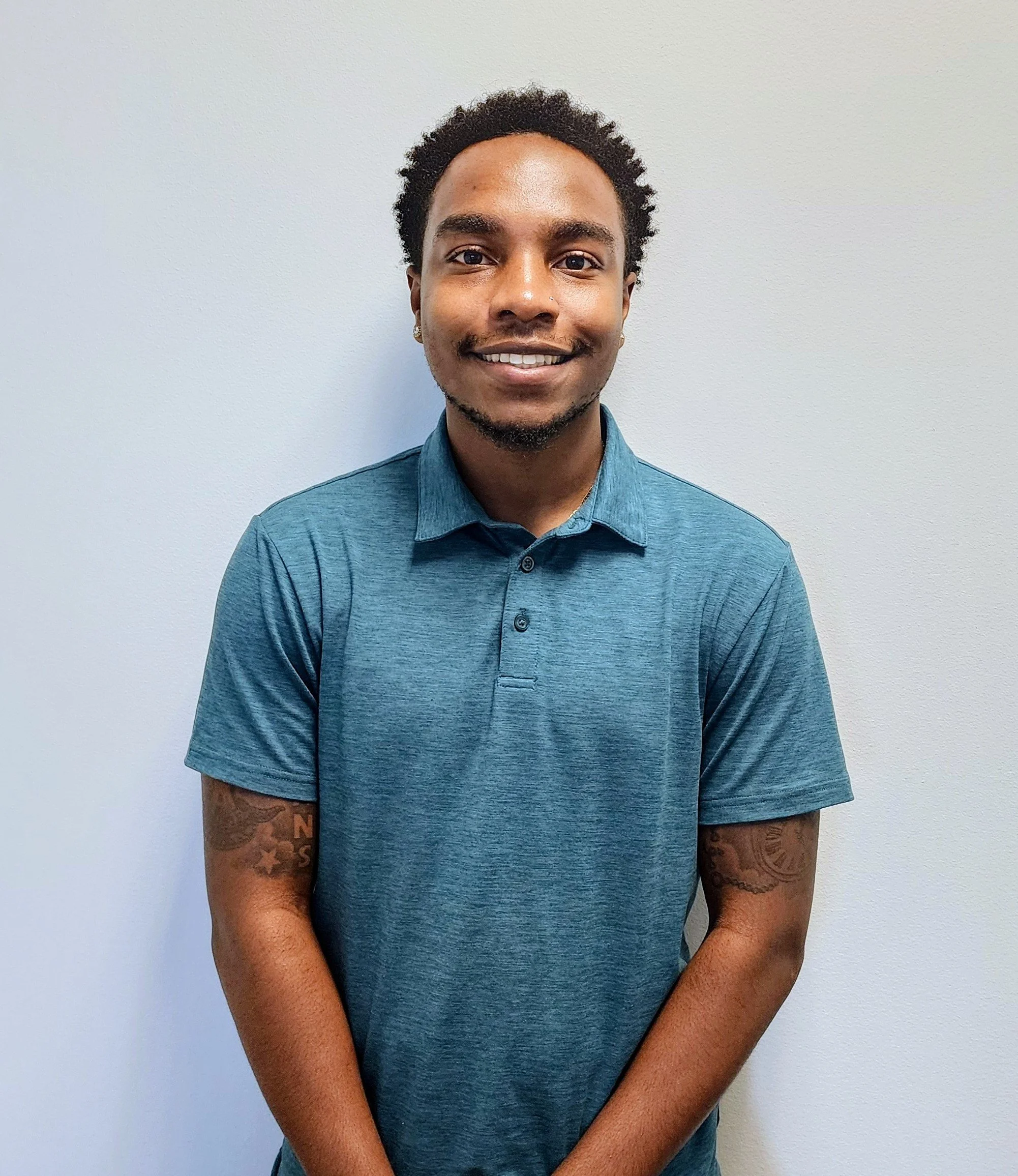 A young man with curly black hair, earrings, and a short beard, smiling, wearing a blue button-up shirt, standing against a plain white wall.