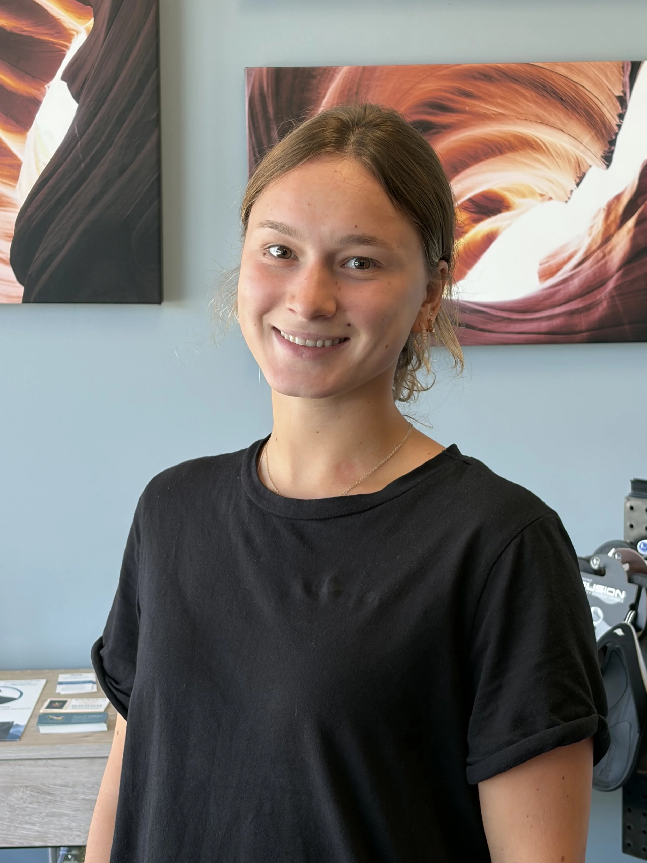 A young woman with short brown hair smiling indoors with two desert canyon landscape photographs on the wall behind her.