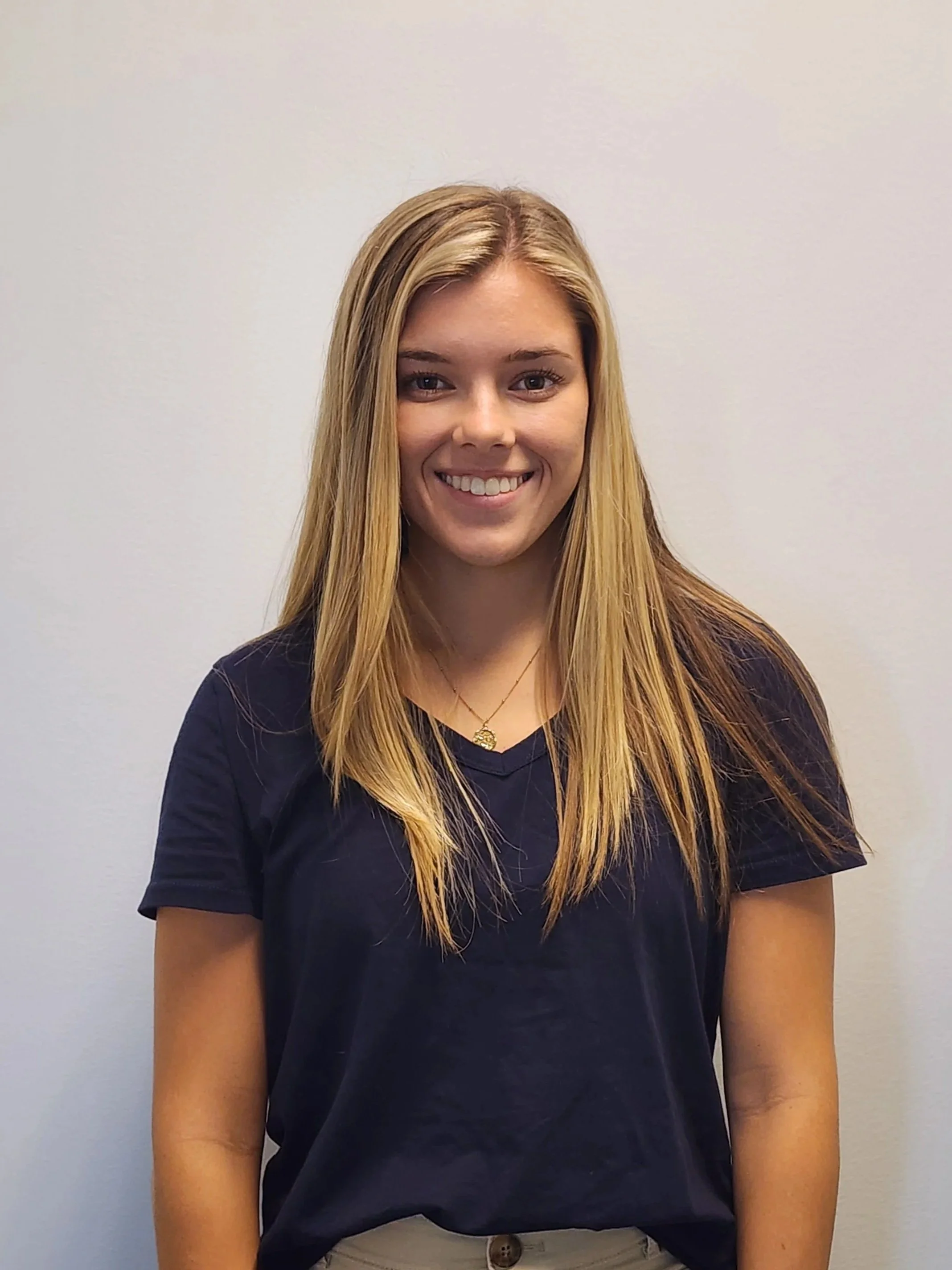 A young woman with long blonde hair smiling, wearing a black t-shirt, a gold necklace, standing against a plain light-colored wall.