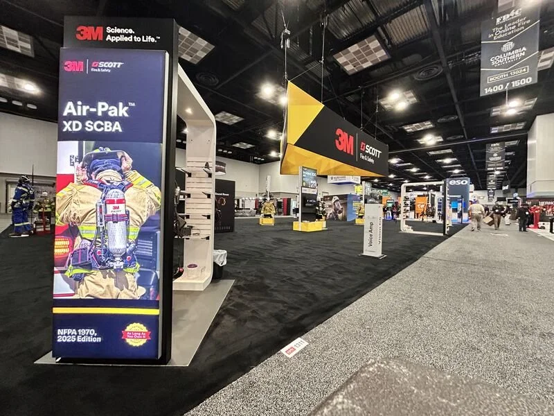 Trade show booth showcasing Scott Air-Pak SCBA breathing apparatus, with signage of 3M and Scott brands, and people walking in the background.