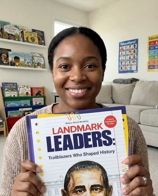 A woman smiling and holding a book titled 'Landmark Leaders: Trailblazers Who Shaped History' in a cozy reading room with bookshelves and posters.
