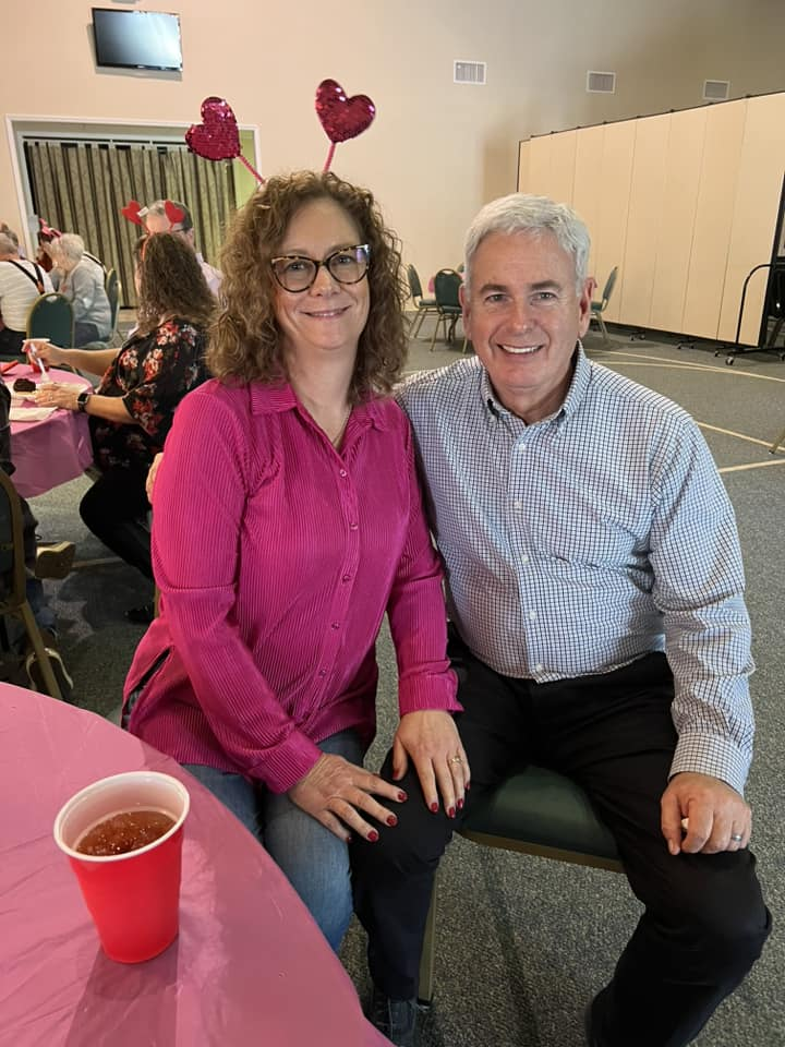 A woman with curly hair wearing glasses and a pink shirt, and a man with gray hair wearing a checkered shirt, sitting close together and smiling at the camera at a celebratory event with pink decorations and a pink tablecloth, with other people in the background.
