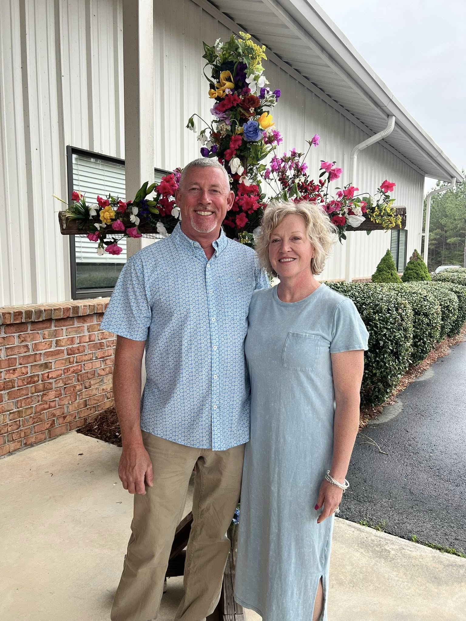A smiling man and woman standing outdoors in front of a flower arrangement and a building with beige siding. The man is wearing a light blue patterned shirt and khaki pants, the woman is wearing a light blue dress. There are green bushes and trees in the background.