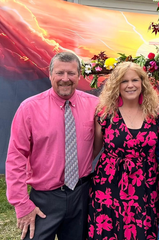 A man and woman standing outdoors in front of a colorful sunset mural with flowers above their shoulders. The man is wearing a pink shirt and a patterned tie, and the woman is dressed in a black dress with a pink floral pattern and large pink earrings.