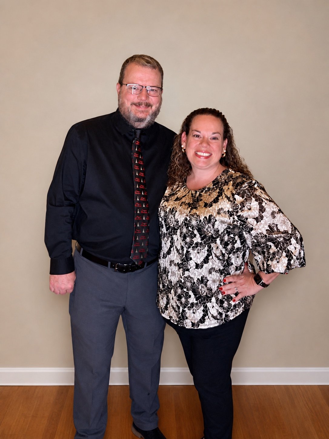 A man with glasses and a beard wearing a black shirt, gray pants, and a festive tie, standing next to a woman with curly hair in a floral-patterned blouse and black pants, both smiling and posing against a plain beige wall.