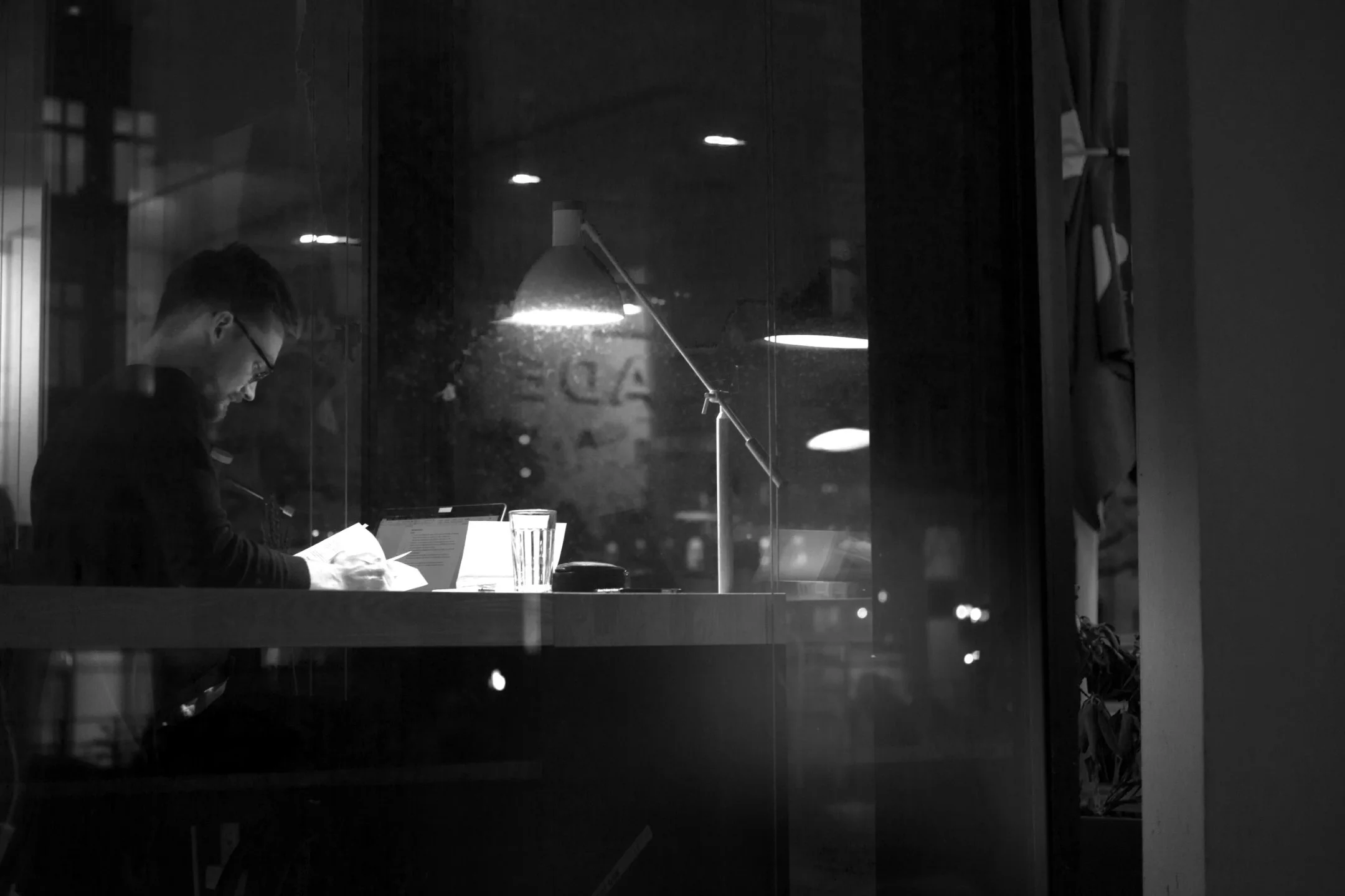 A man with glasses and a beard is sitting at a table in a dimly lit room, reading a book. There is a glass of water in front of him, and the room has a lamp and large windows with reflections of lights and the street outside.