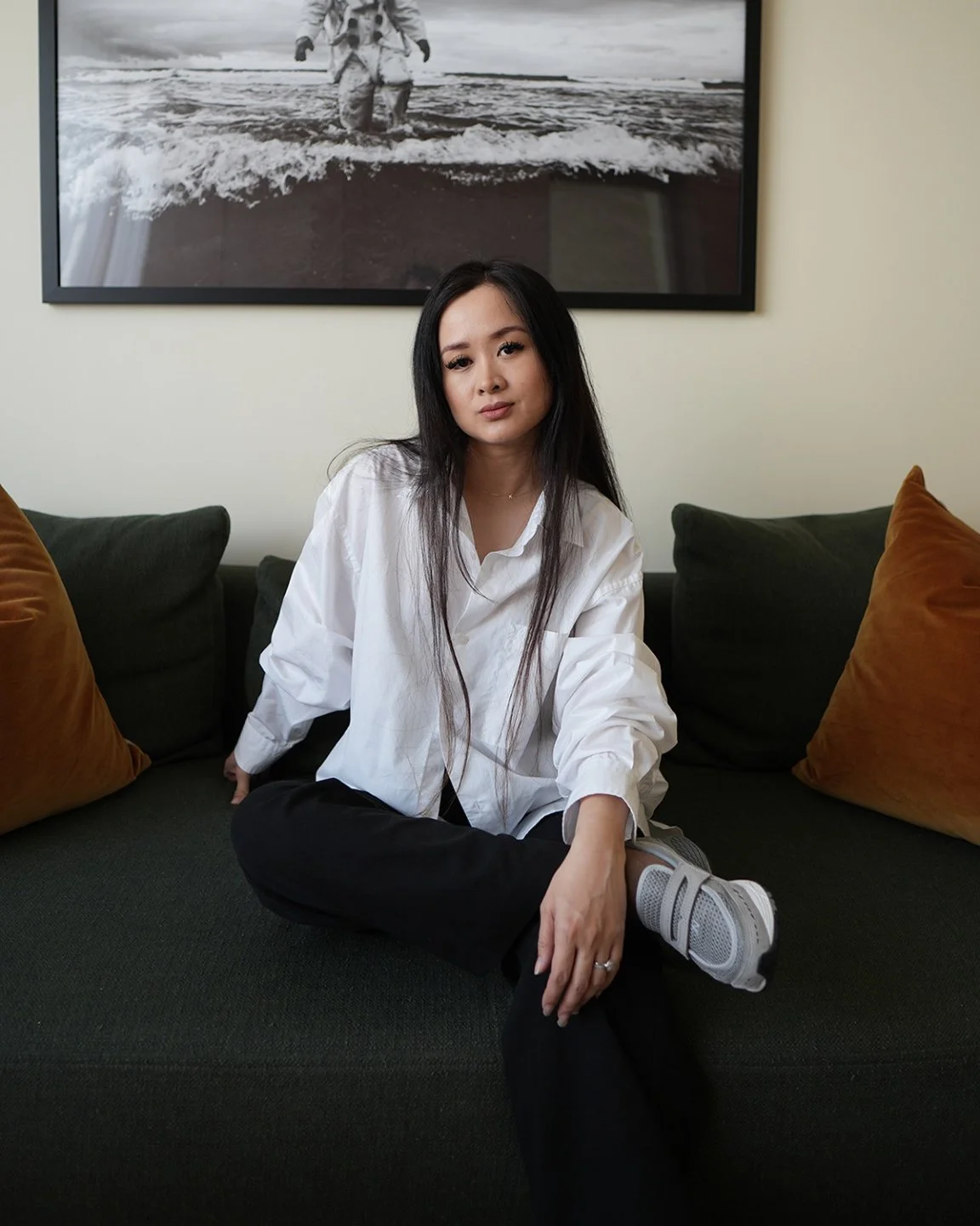 A young woman with long black hair sitting on a dark green couch with orange pillows, wearing a white button-up shirt, with a large black and white photograph of a person walking in the water hanging behind her.