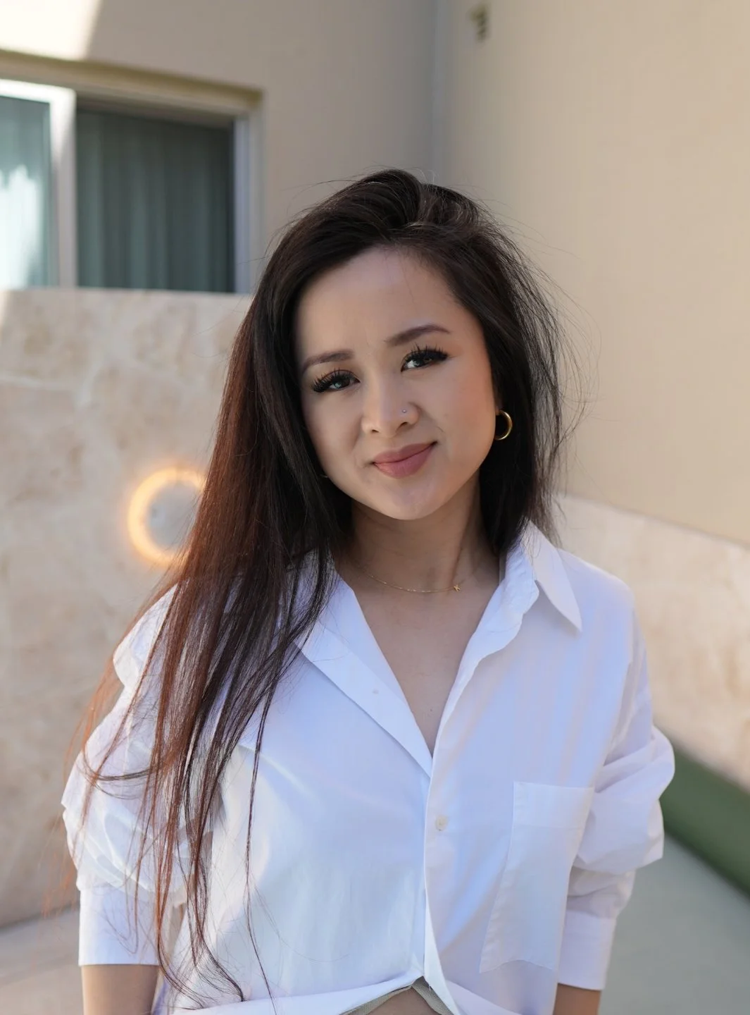 A woman with long dark hair, wearing a white shirt and gold hoop earrings, smiling and looking at the camera.