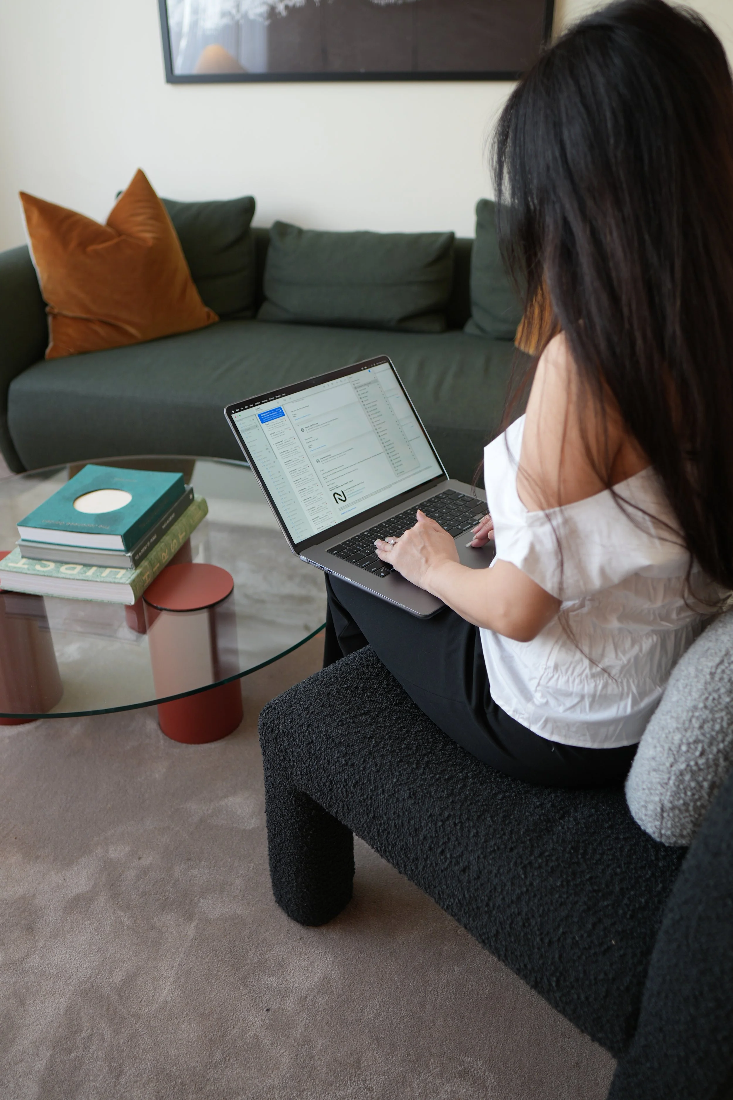 A woman with dark hair is sitting on a couch, working on a laptop placed on her lap in a living room. There are stacked books on the glass coffee table in front of her and a green couch with a brown pillow in the background.