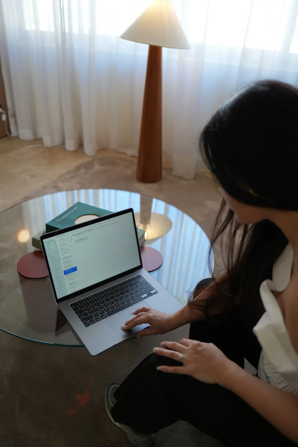 Woman working on a laptop at a glass coffee table in a bright room with sheer white curtains, a wooden floor, and a floor lamp.