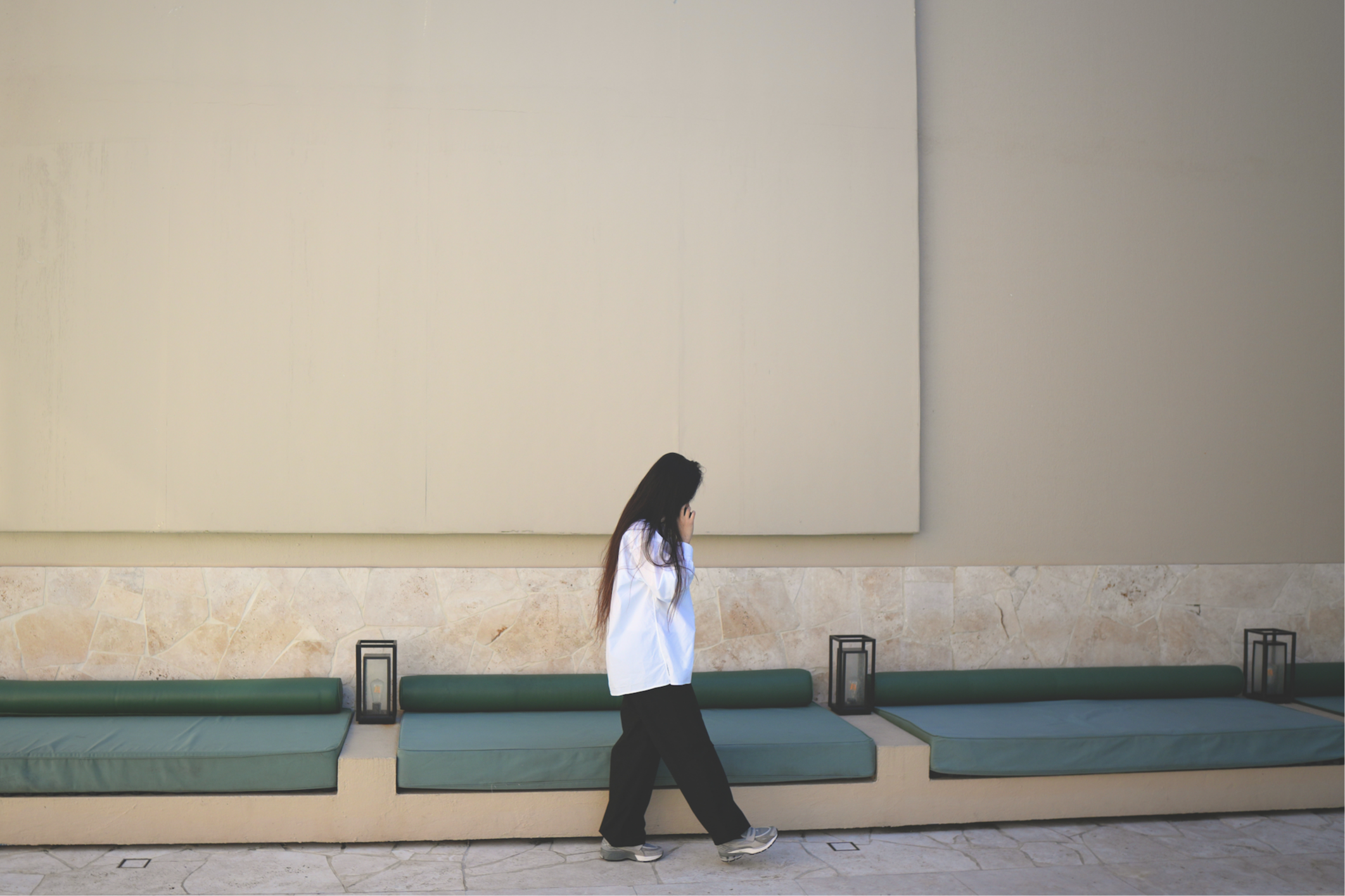 A woman walking past a wall with three green cushioned benches and black lanterns. The wall has a stone base and a large plain panel above.