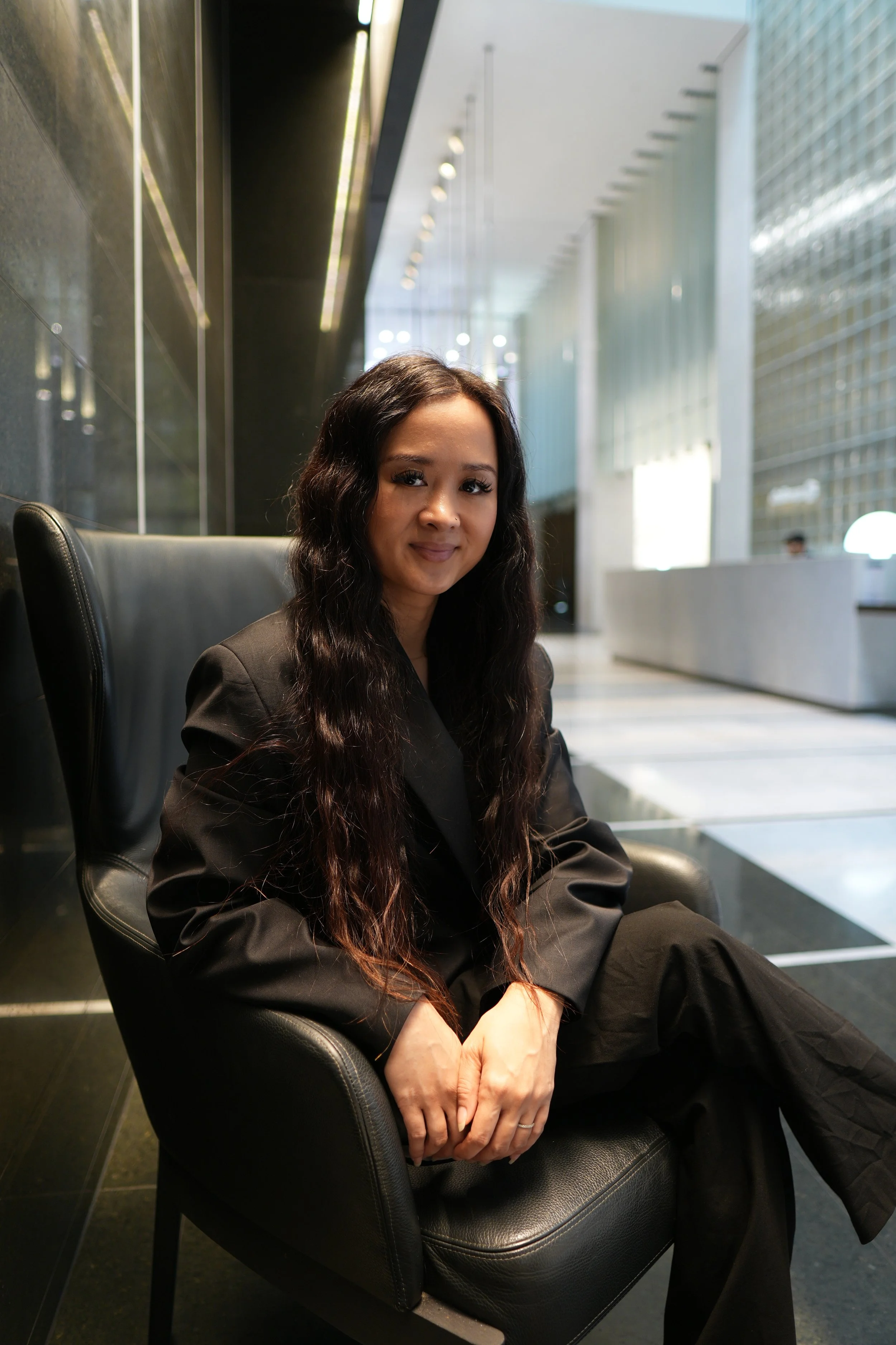 A woman with long dark hair sitting in a black leather chair in a modern, bright lobby area with large windows and contemporary design elements.
