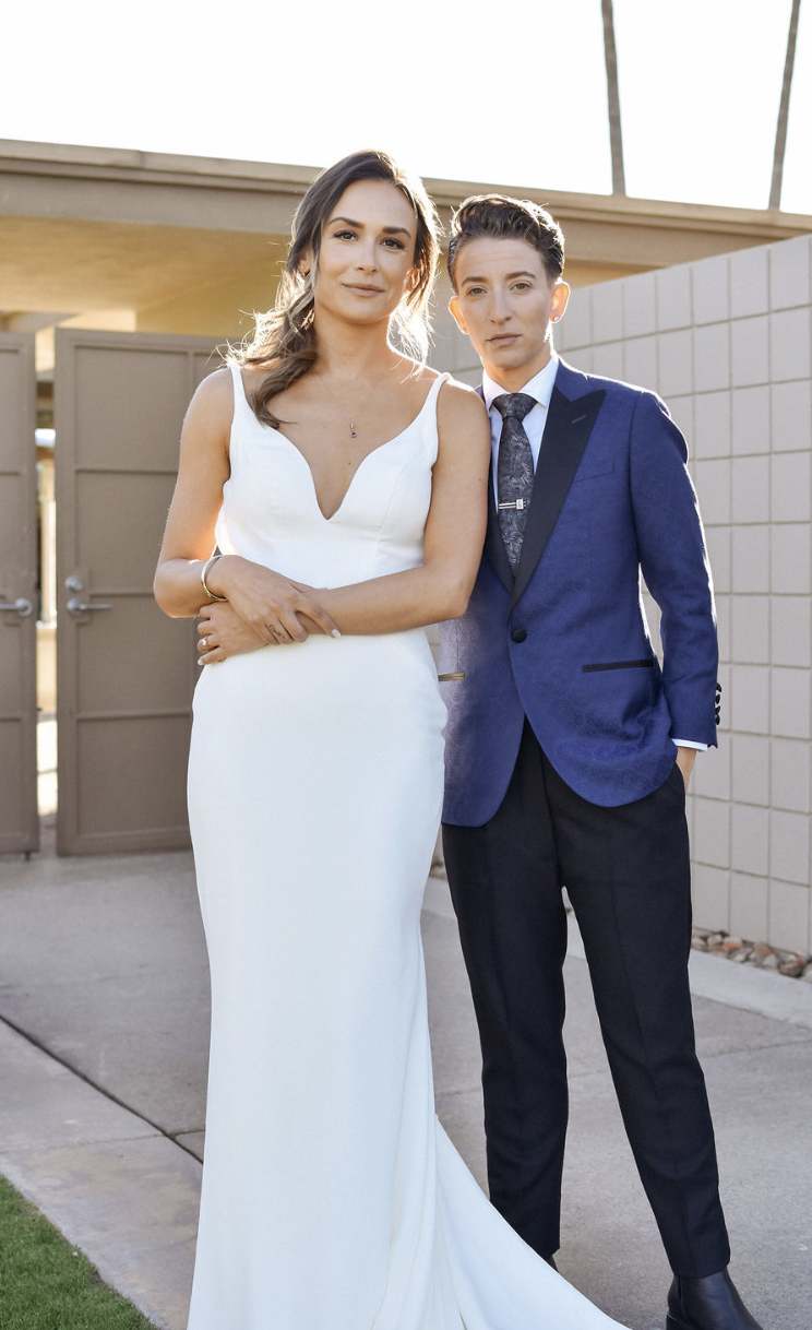 Two women standing together outdoors, one in a white dress and the other in a blue suit, with a beige building and palm trees in the background.