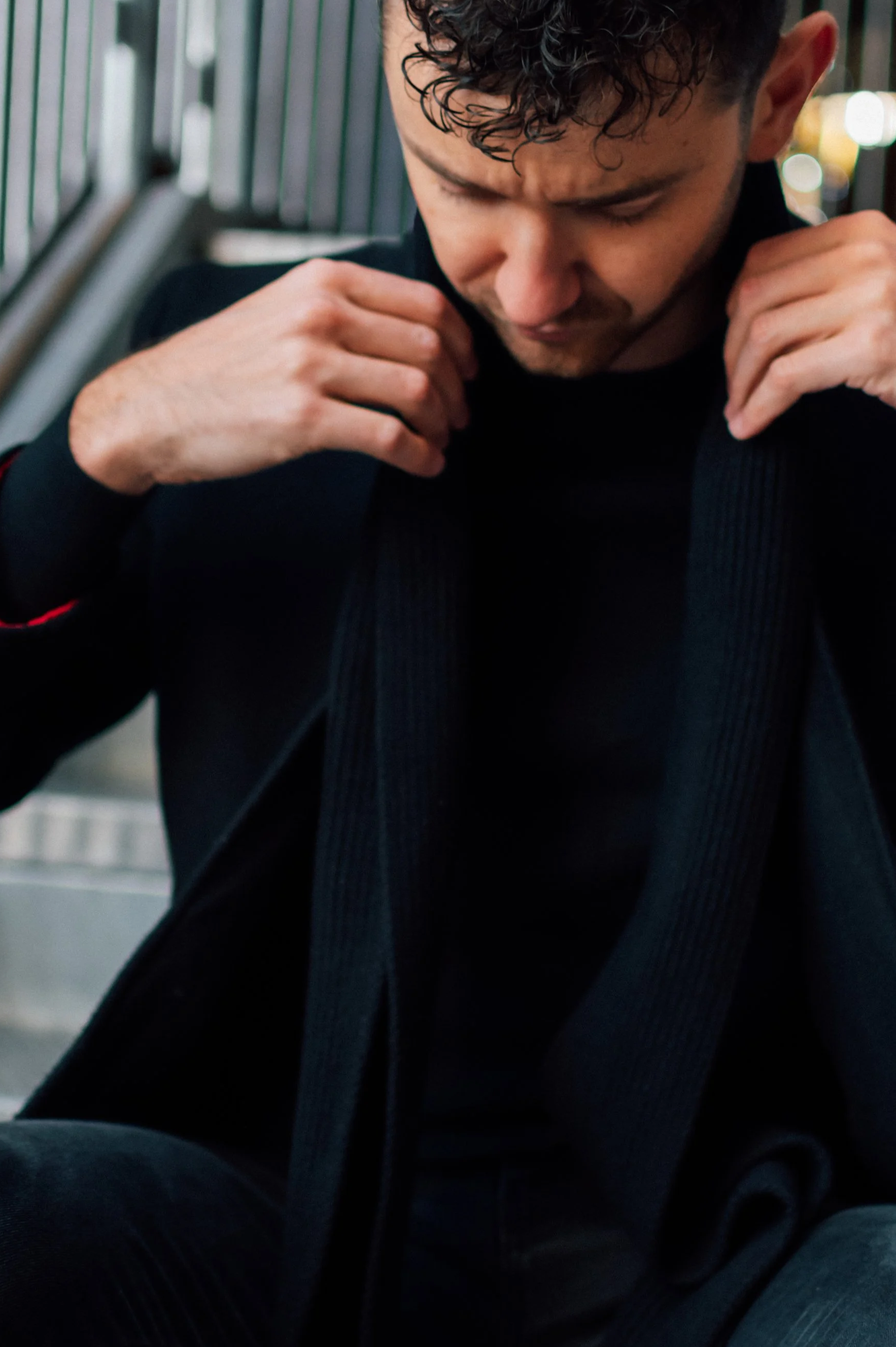 A young man with curly dark hair sitting on stairs, adjusting his black jacket with a somber expression.