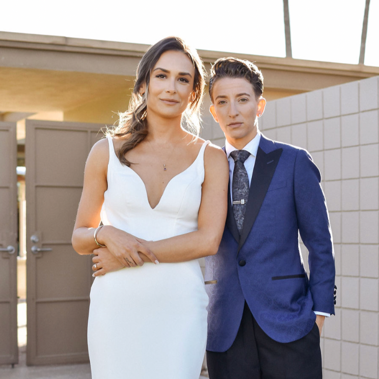 A woman in a white dress standing next to a woman in a blue suit, both outside near a brick wall and a tan-colored structure.