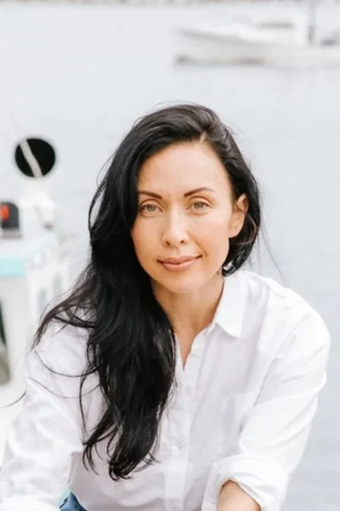 Woman with long dark hair wearing a white shirt near a body of water with boats in the background.