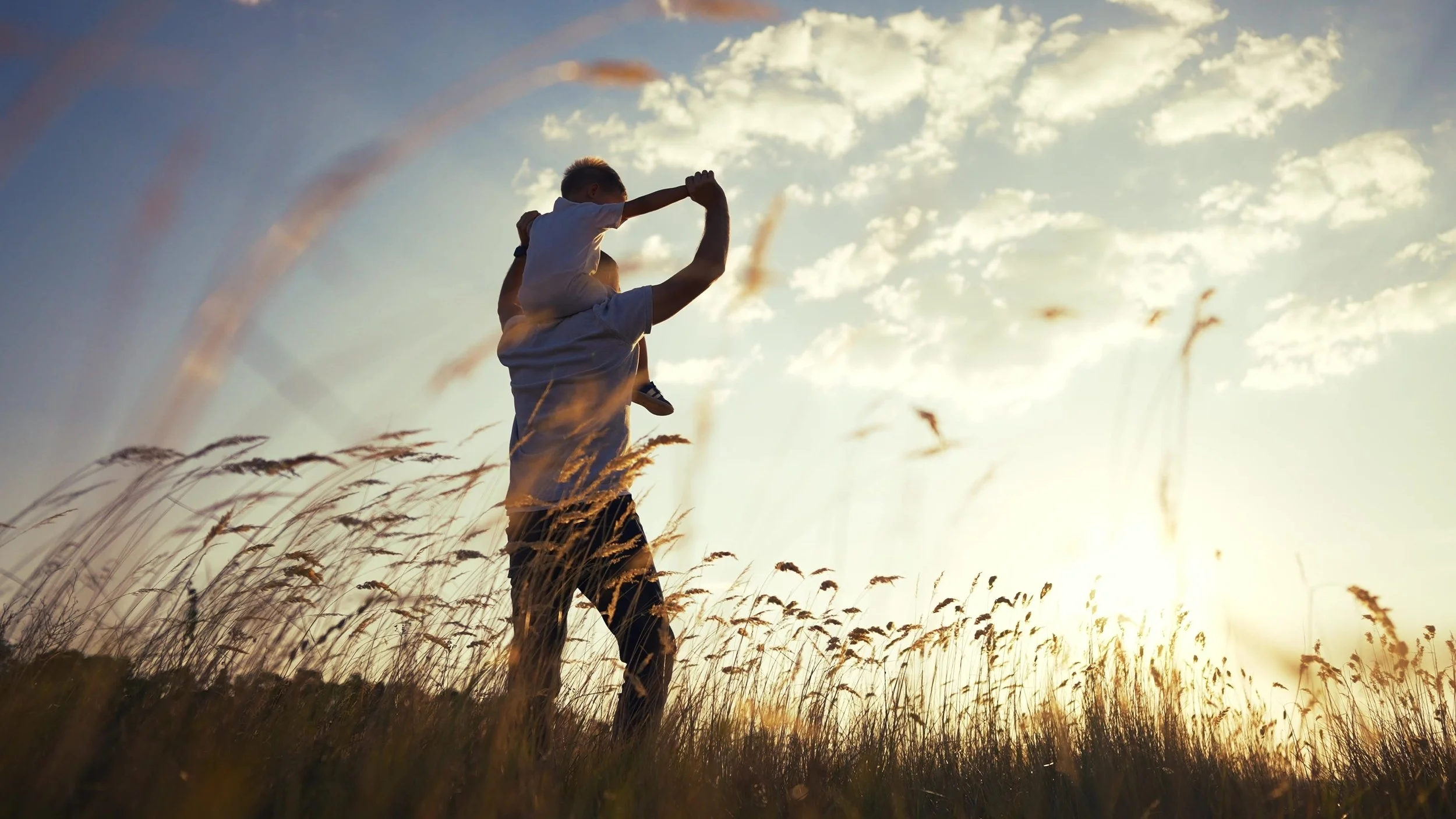 A young boy sits atop his father's shoulders. Facing away from the camera, they enjoy the sunset as it passes over a prairie.