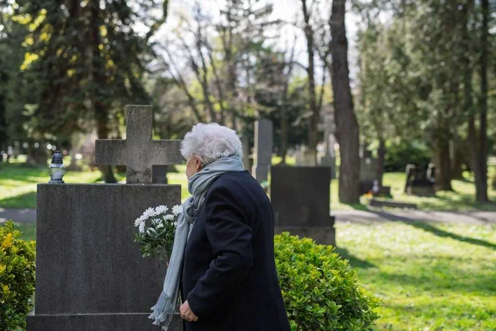 An elderly woman in a black coat places white flowers atop a loved one's grave.