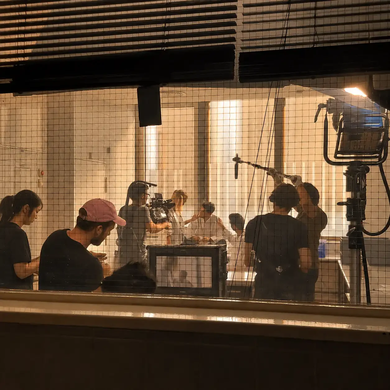 Behind-the-scenes view of a film or television production crew filming a scene in a kitchen with sunlight streaming through large windows. Crew members are operating cameras and equipment, with some conversing and others focused on filming.