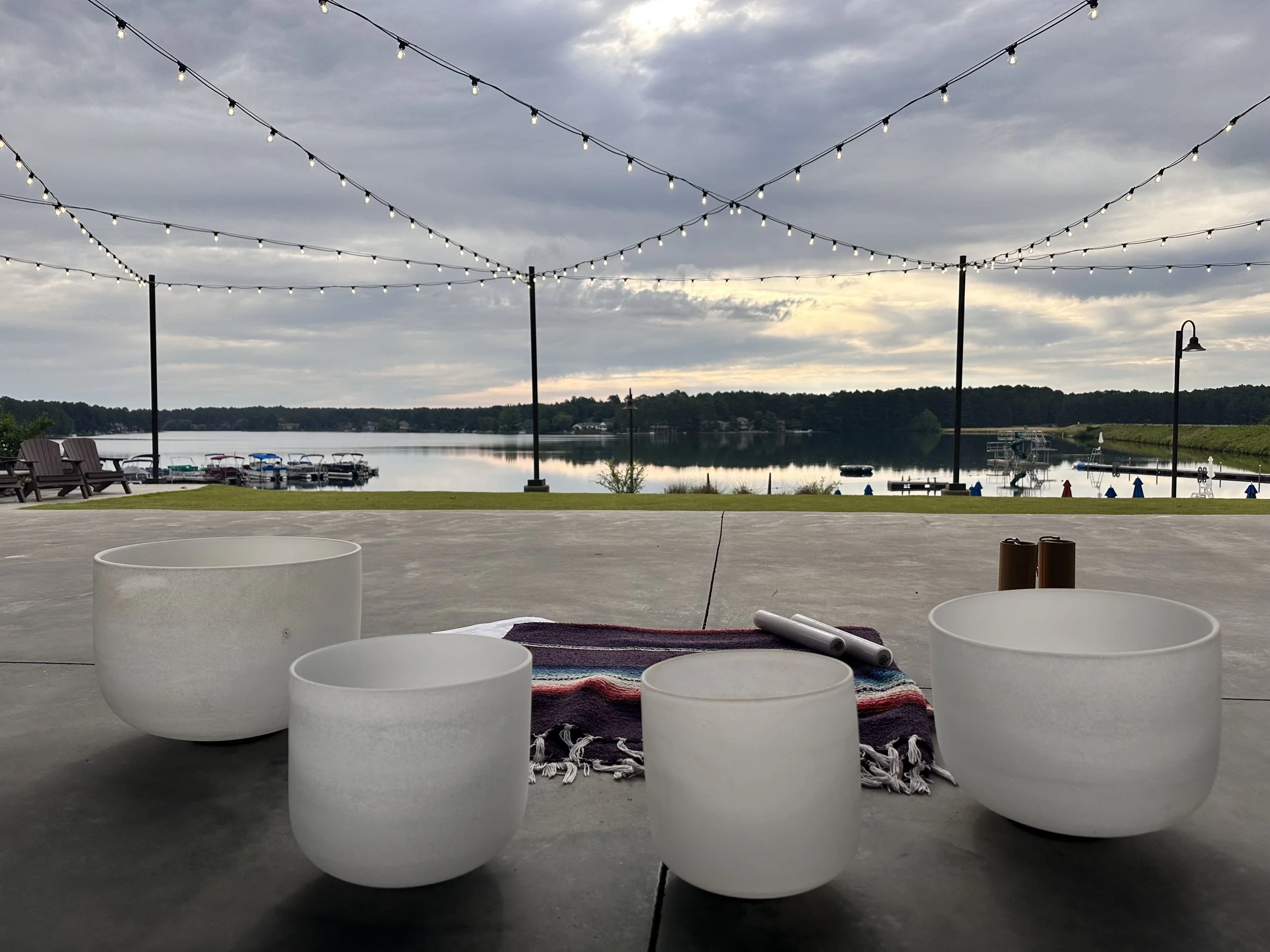 Four white singing bowls surrounded by a woven blanket overlooking a lake with docks, boat, and string lights under an overcast sky.