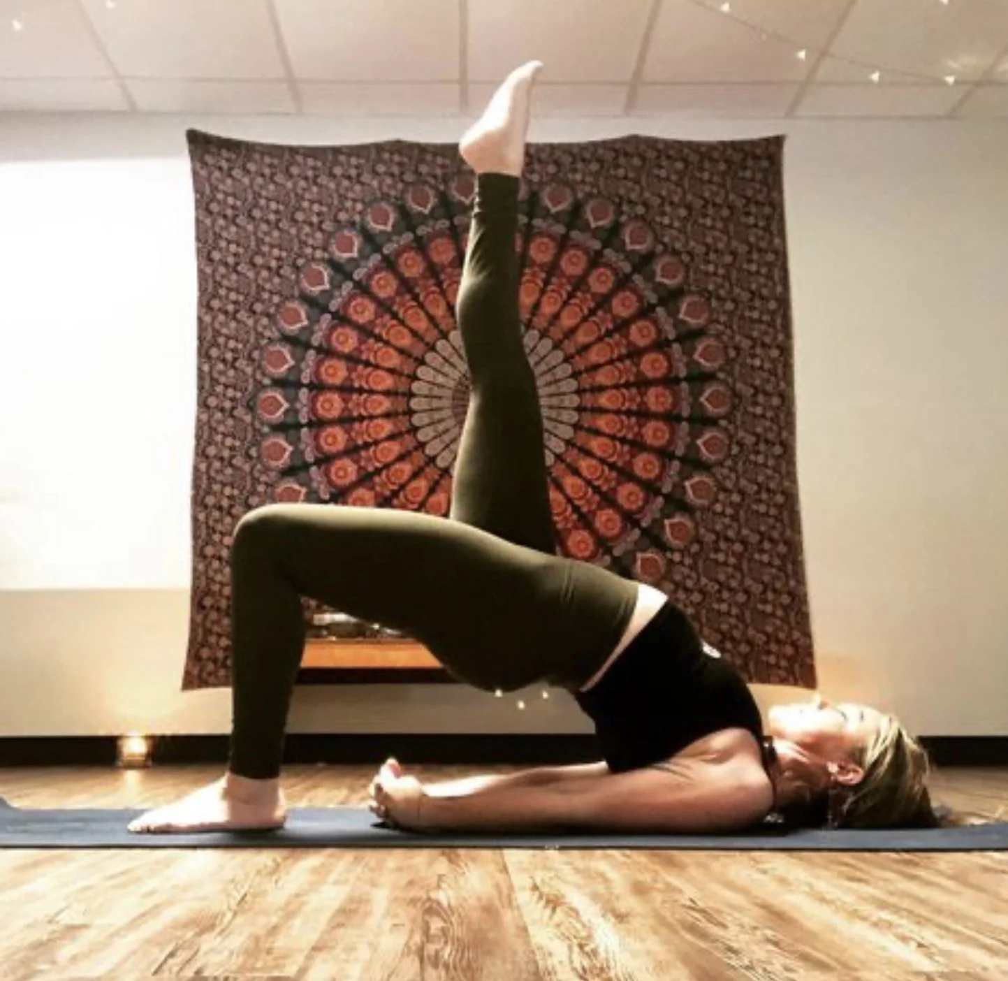 A woman practicing yoga indoors on a yoga mat, performing a pose with one leg extended vertically and head and shoulders on the floor in a bridge position, with a decorative mandala tapestry hanging on the wall behind her.