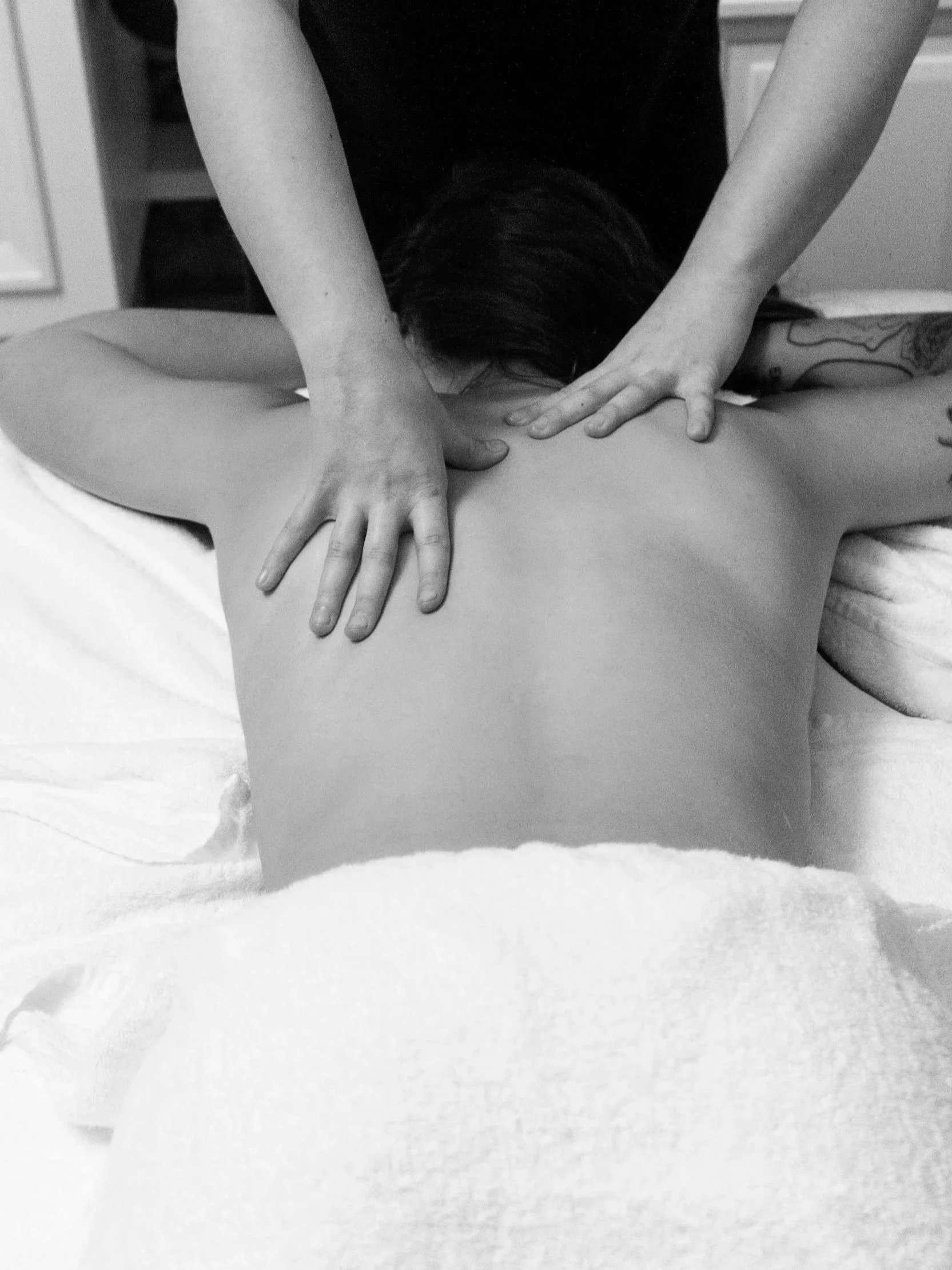 Black and white photo of a person receiving a massage, lying face down on a massage table, with two hands applying pressure to their back.