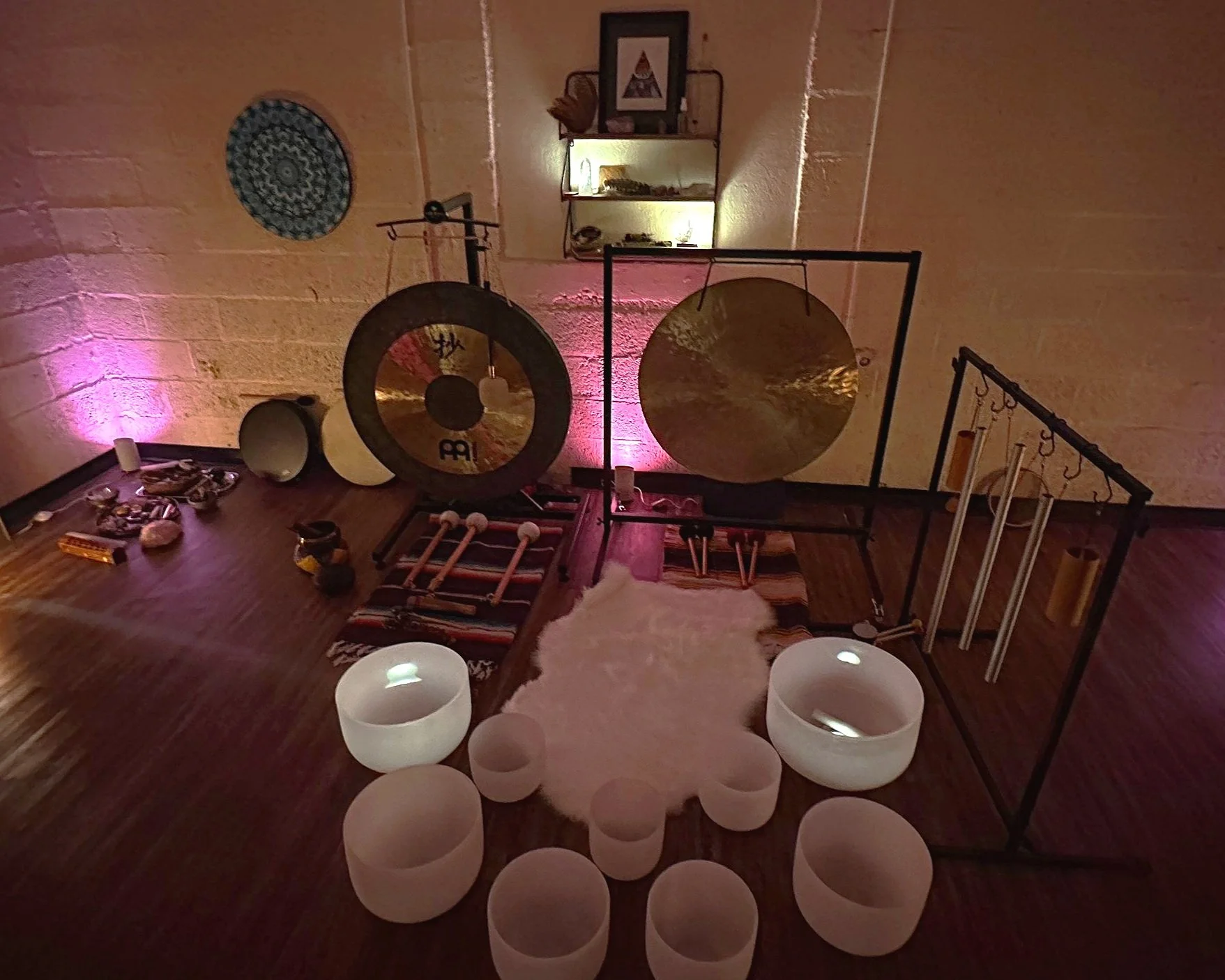 A room decorated with musical instruments and meditation supplies, including gongs, crystal bowls, Tibetan bowls, chimes, and candles, with soft pink lighting.