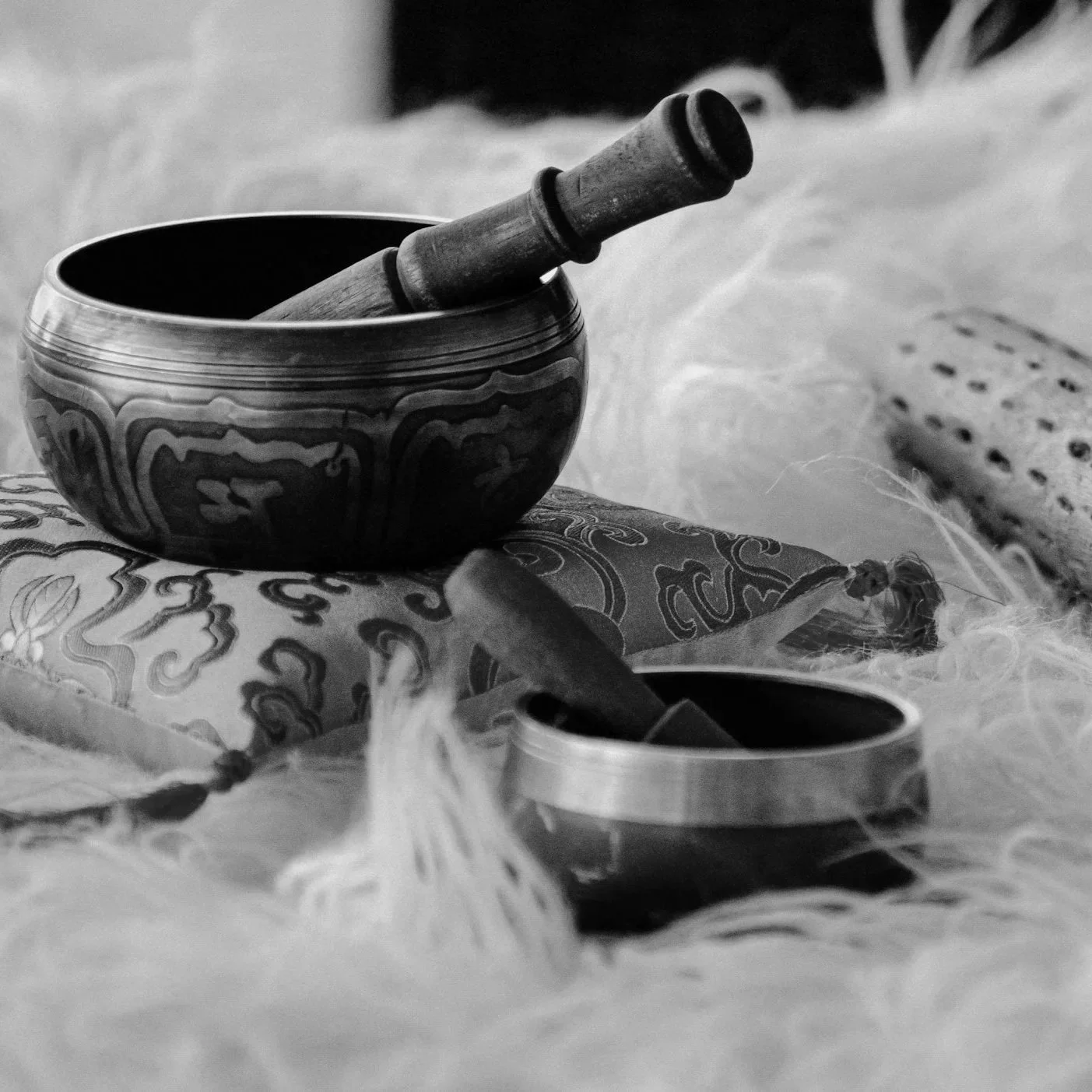 A black and white photo features a Tibetan singing bowl with a mallet resting inside, placed on embroidered fabric, with a small metal container and another mallet nearby, all surrounded by soft feathers.