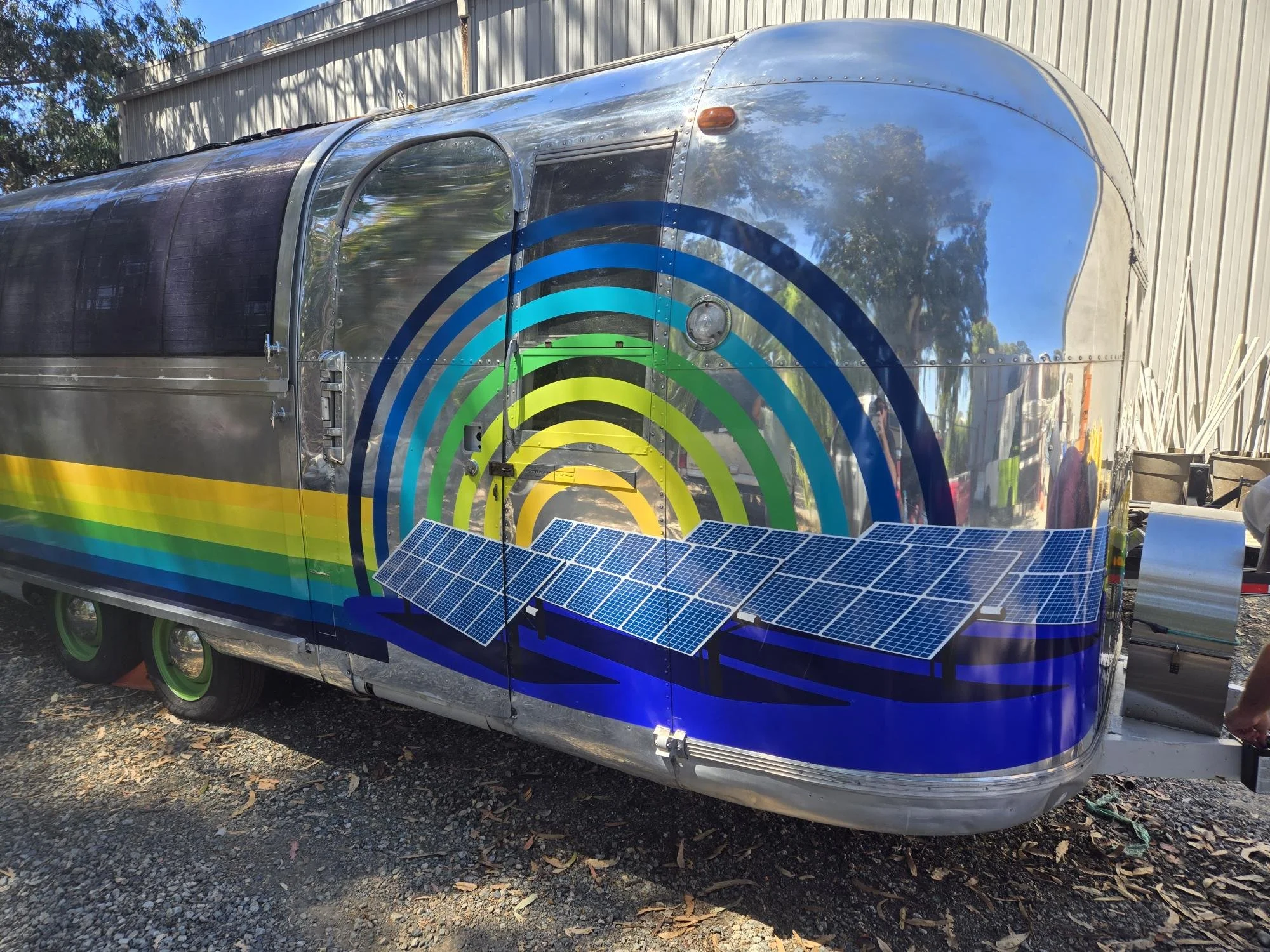 A photo of a silver Airstream trailer with custom graphics. They feature a yellow, green, and blue stripe along the side, and solar panels on a purple field, with rings of yellow, green, and blue behind them like a rising sun.