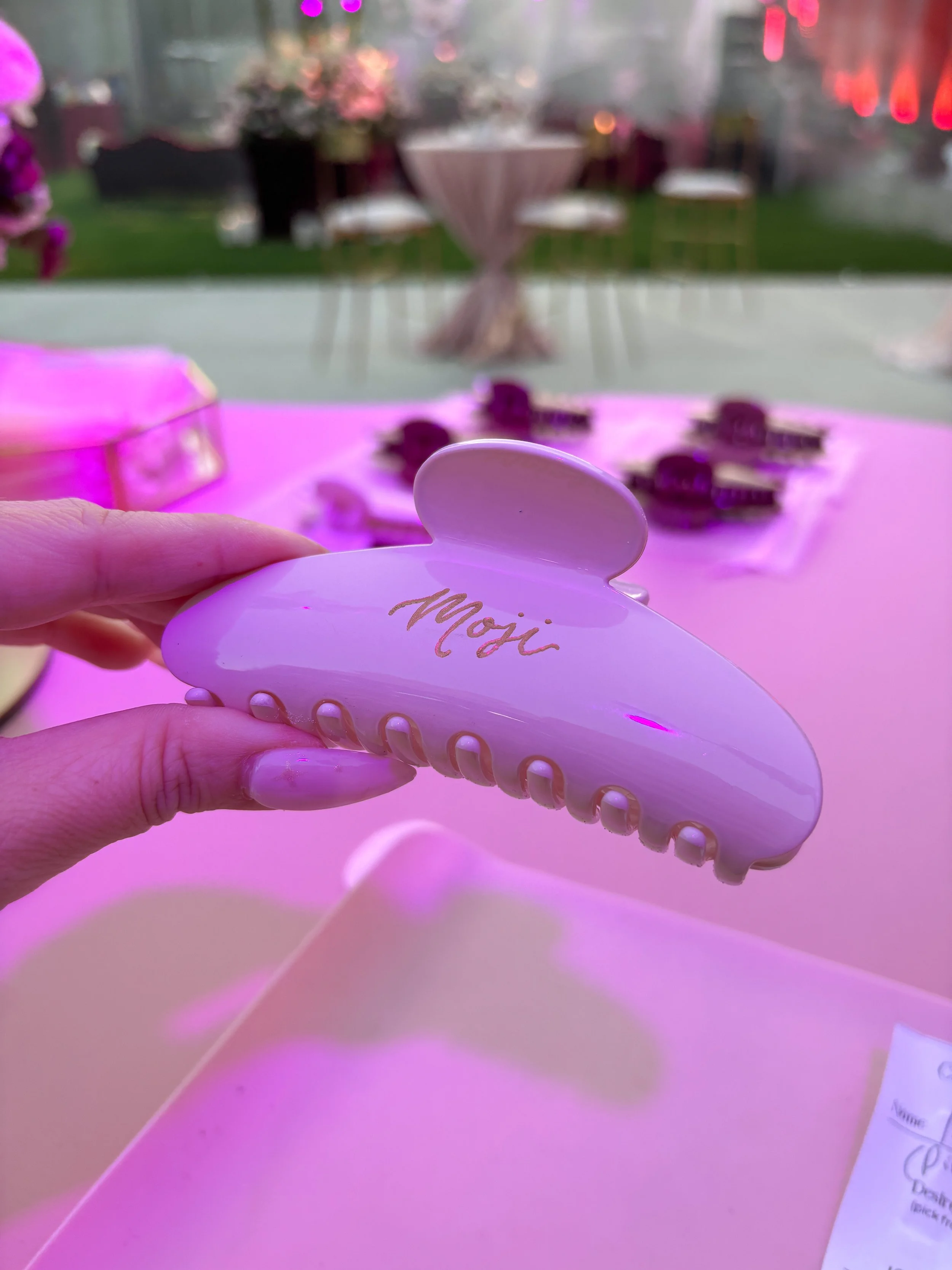 A hand with long pink nails holding a white claw clip with the name "Moji" engraved on it and filled with gold paint