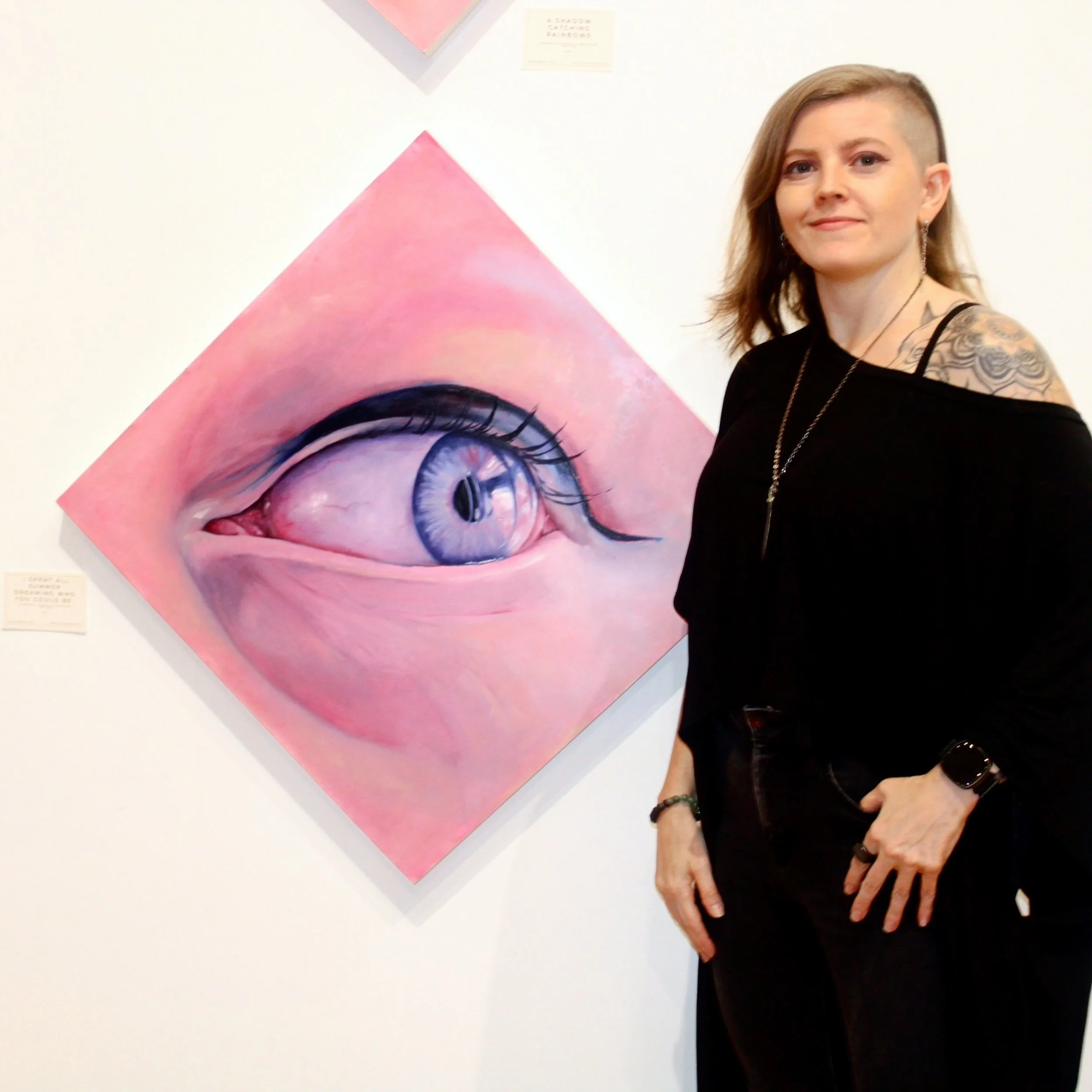 A woman with a blonde, shaved hairstyle and tattoos on her shoulder is standing next to a large, colorful painting of a woman's eye with prominent eyelashes on a wall in an art gallery.