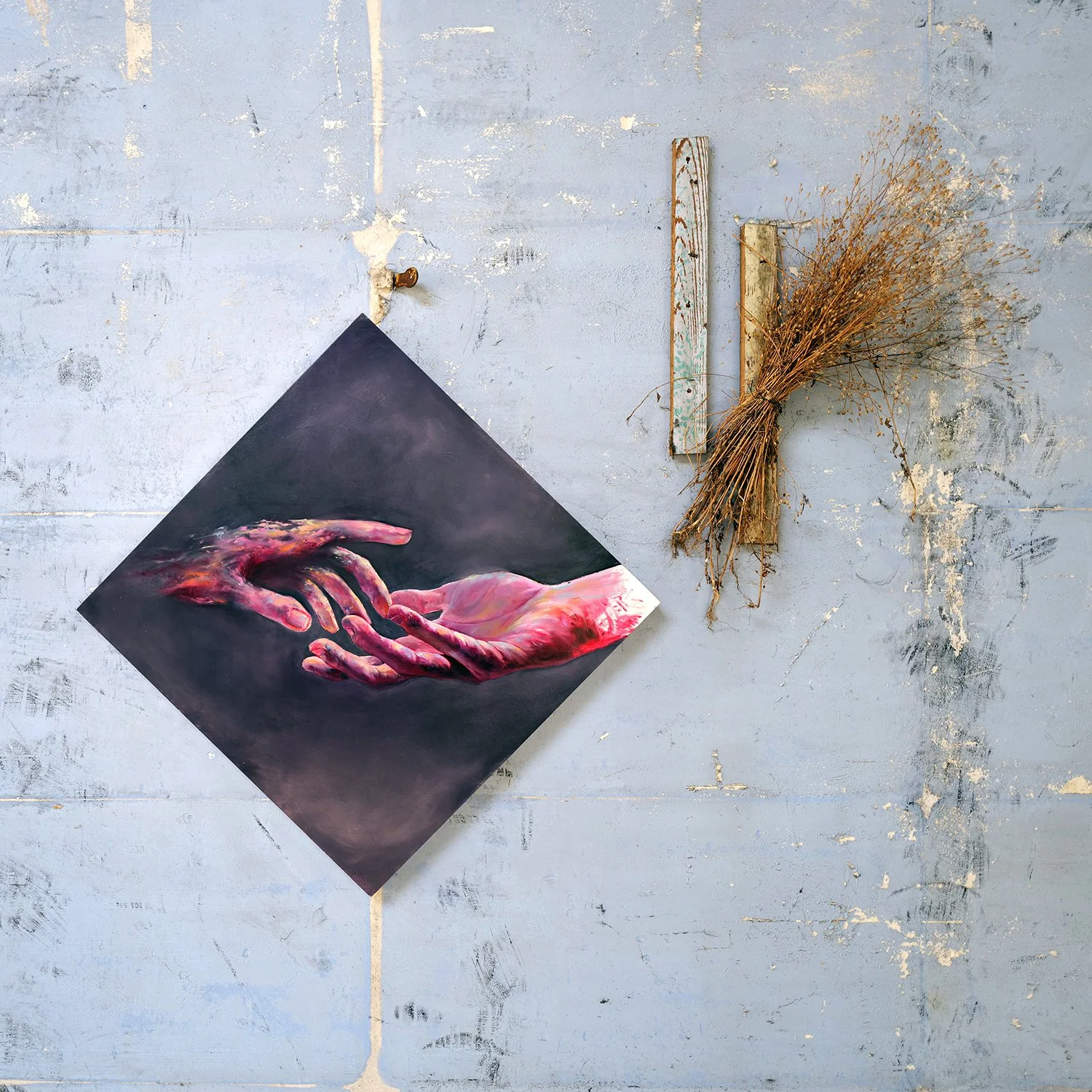 A wall with peeling paint, a painting of two hands reaching towards each other, and a bundle of dried twigs attached to the wall.