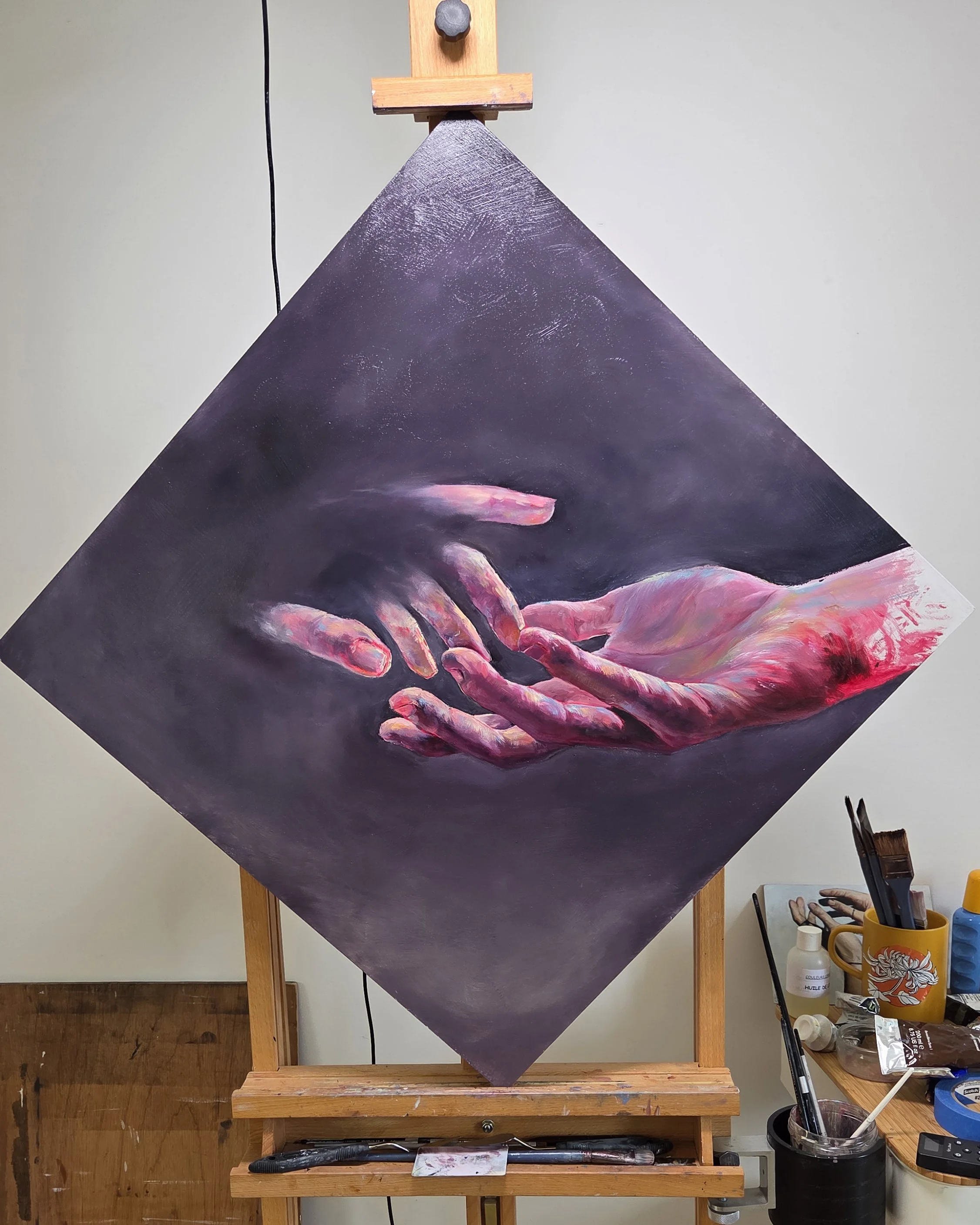 A painting of two hands reaching towards each other. The workspace includes brushes, paints, and art supplies.