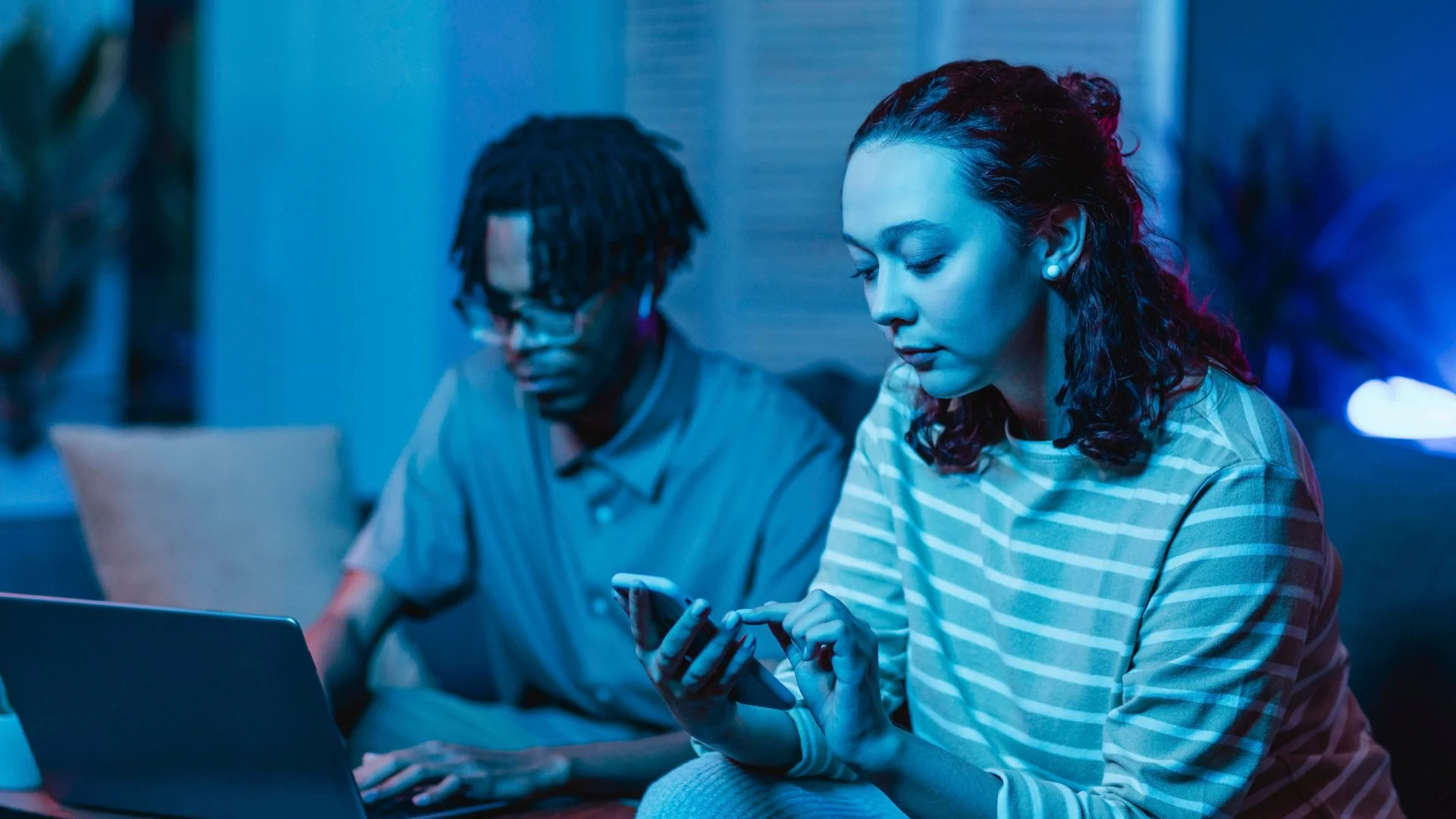 A woman and a man sitting on a couch in a dimly lit room, both looking at their smartphones. The woman is wearing a striped shirt and has curly hair, while the man has dreadlocks and glasses. A laptop is open in front of them.