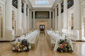 Elegant wedding reception setup inside a grand hall at the State Library Victoria with long white tables, gold chiavari chairs, and lush floral arrangements at each end under a skylit ceiling.