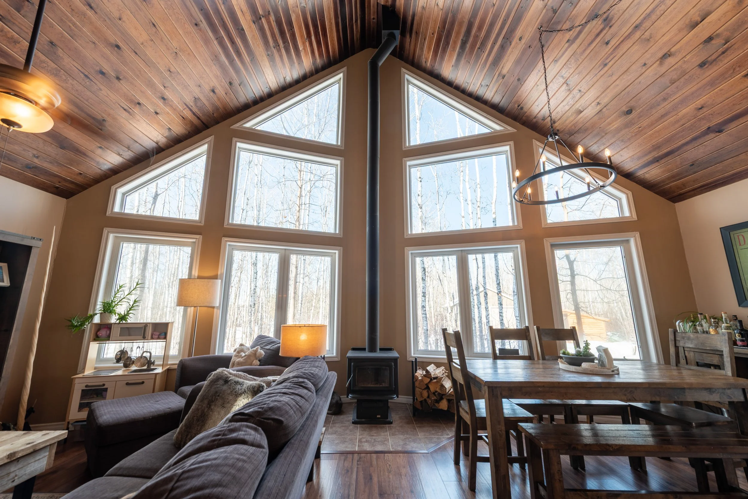 A cozy living room with a high wooden ceiling and large triangular windows letting in natural light, with a wood-burning stove in the center, a sofa, a dining table, and a view of trees outside.