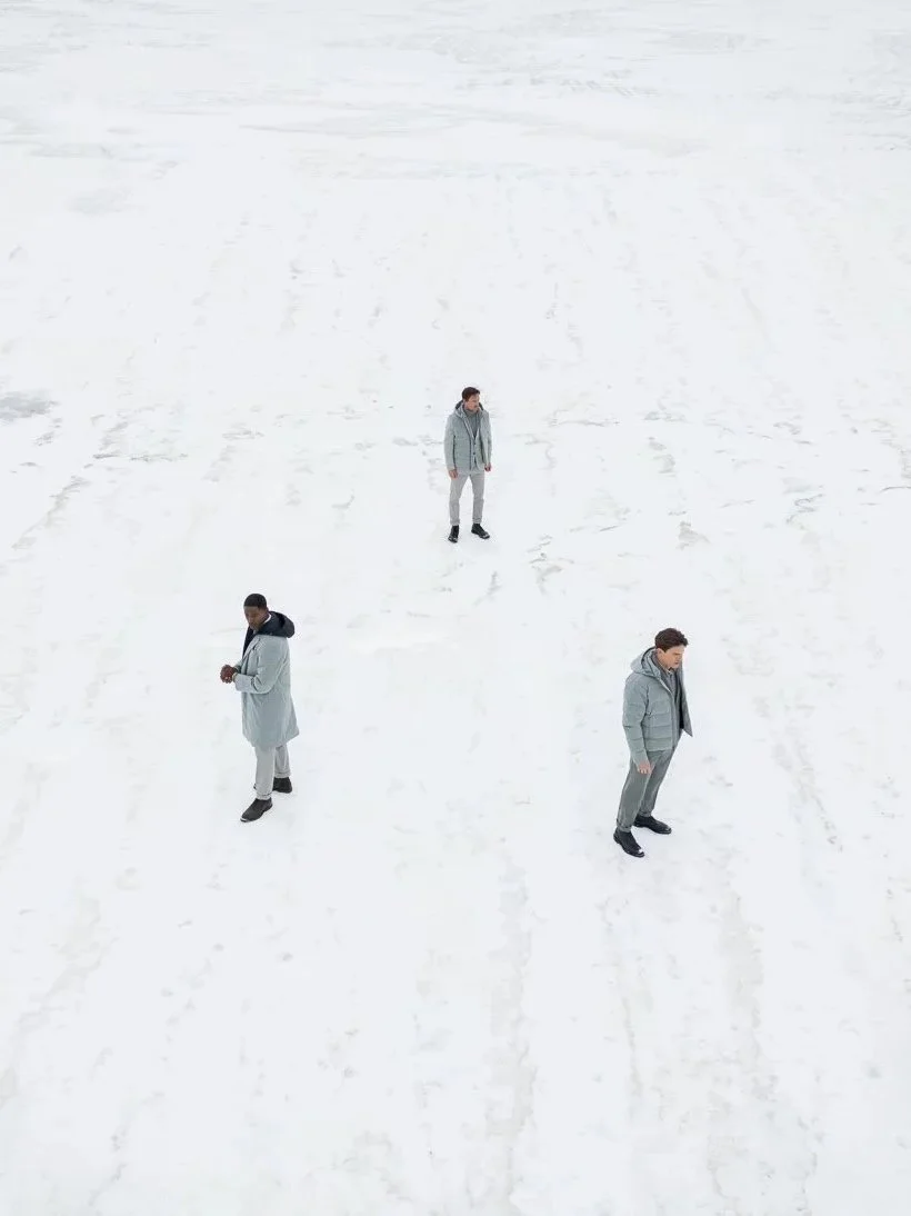 Three men standing on a snowy, icy landscape with tire tracks and footprints visible in the snow.