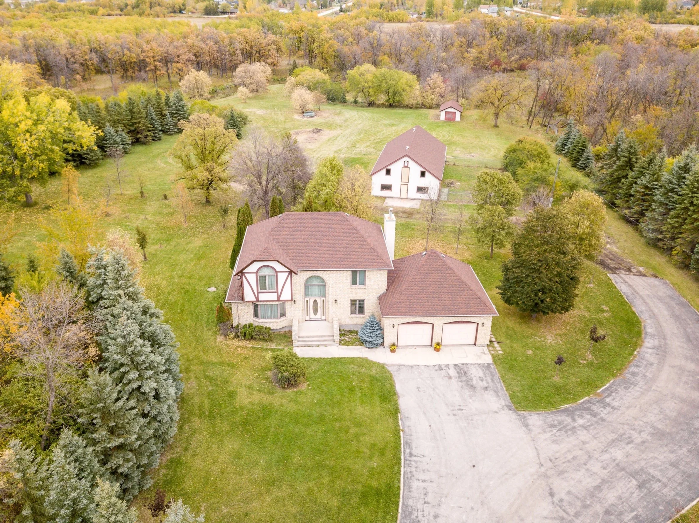 Aerial view of a house with a red roof, surrounded by a lush green lawn, multiple trees, and a winding concrete driveway. There is another smaller building or shed in the background surrounded by trees, and a large wooded area beyond that with more trees and a distant residential area.