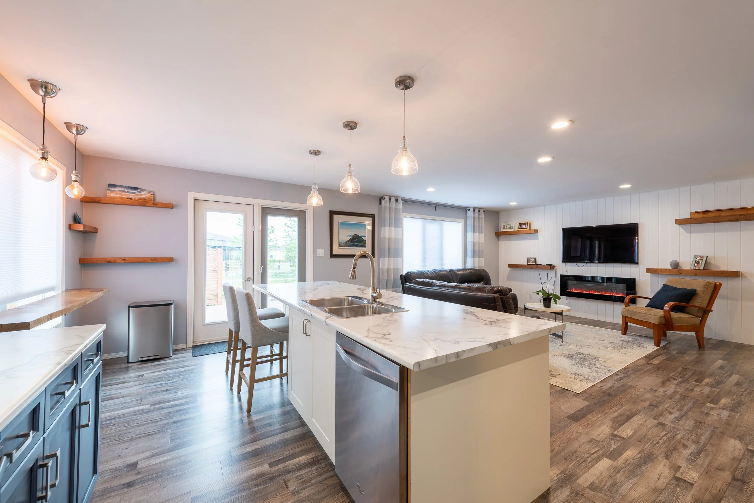 Open-concept living room and kitchen with a white marble island, grey cabinetry, wooden floating shelves, and a wall-mounted TV above an electric fireplace, with large windows and a sliding door bringing in natural light.