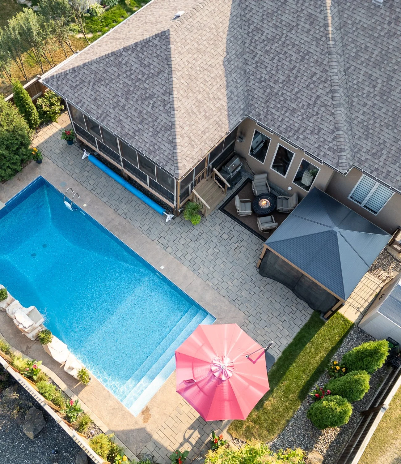 A backyard with a swimming pool, a paved patio area, a house with a screened porch, and outdoor furniture including a fire pit and chairs. There is also a pink umbrella and some plants and trees surrounding the space.